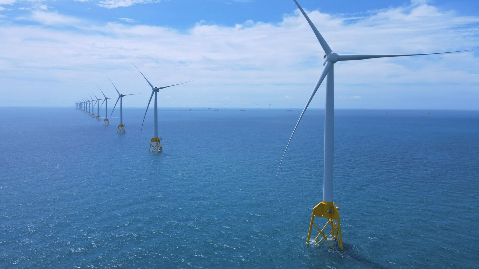 Aerial view looking down a row of three-bladed wind turbines in a calm blue sea, extending into the distance right to left