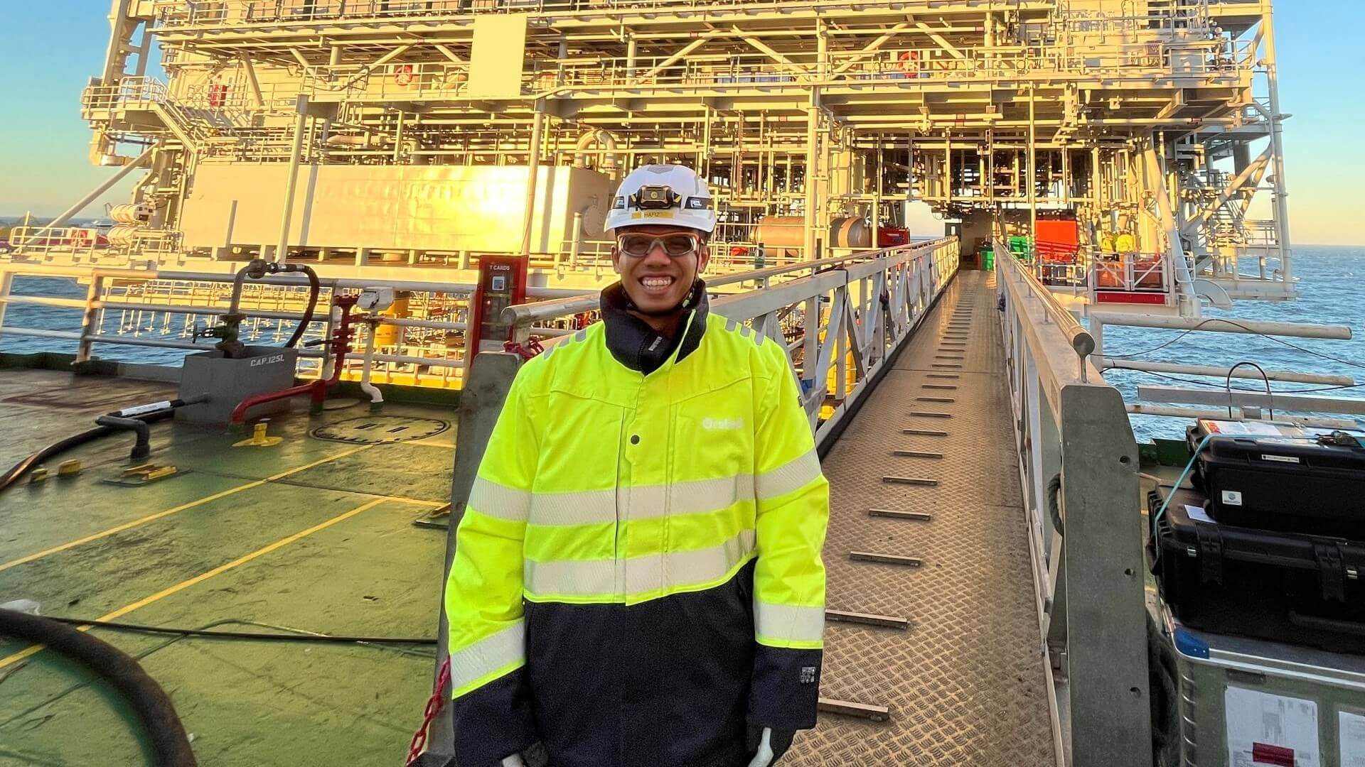 Photo of person in yellow high-viz jacket and hard hat standing on walkway leading to large industrial platform, with lots of pipework and equipment, standing in the sea