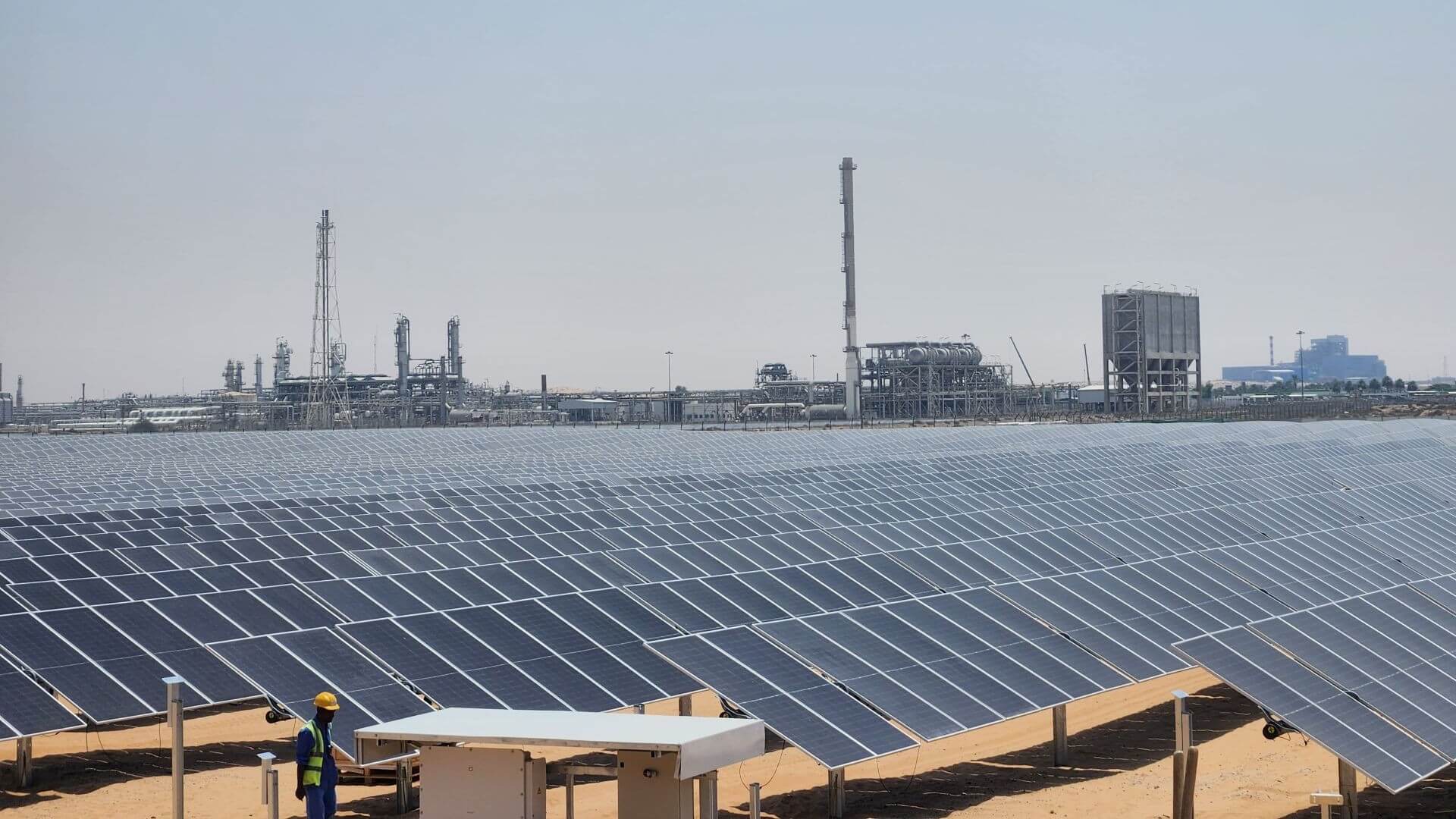 Rows of solar panels in foreground, with industrial plant buildings on the horizon in background