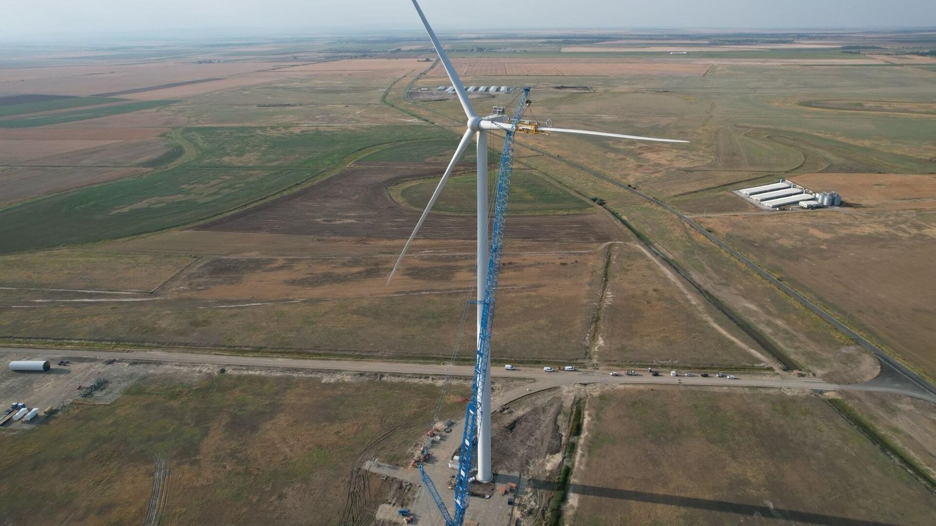 Aerial view of a crane attaching the third blade to a wind turbine being built in green field