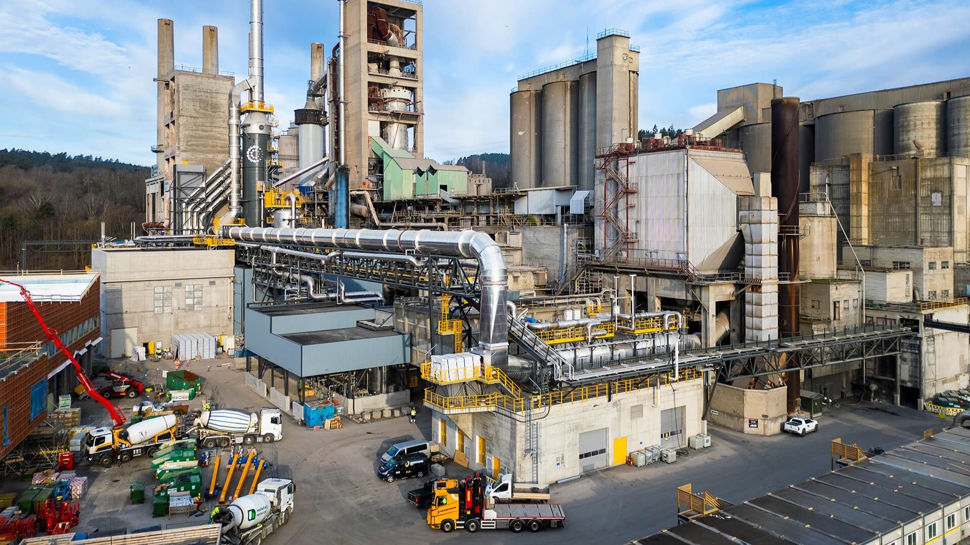 Large industrial plant comprising many grey concrete block buildings with shiny metal pipework in the foreground. Various trucks, cement mixers and cars in front of plant.