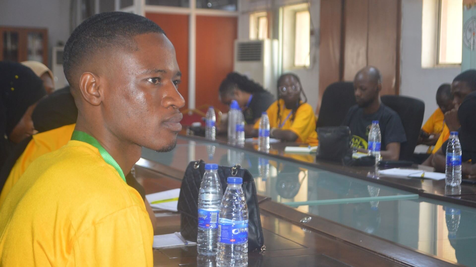 Close up of a student in bright yellow t-shirt, sat at a long desk, with a row of other students sat along a long desk opposite in the background