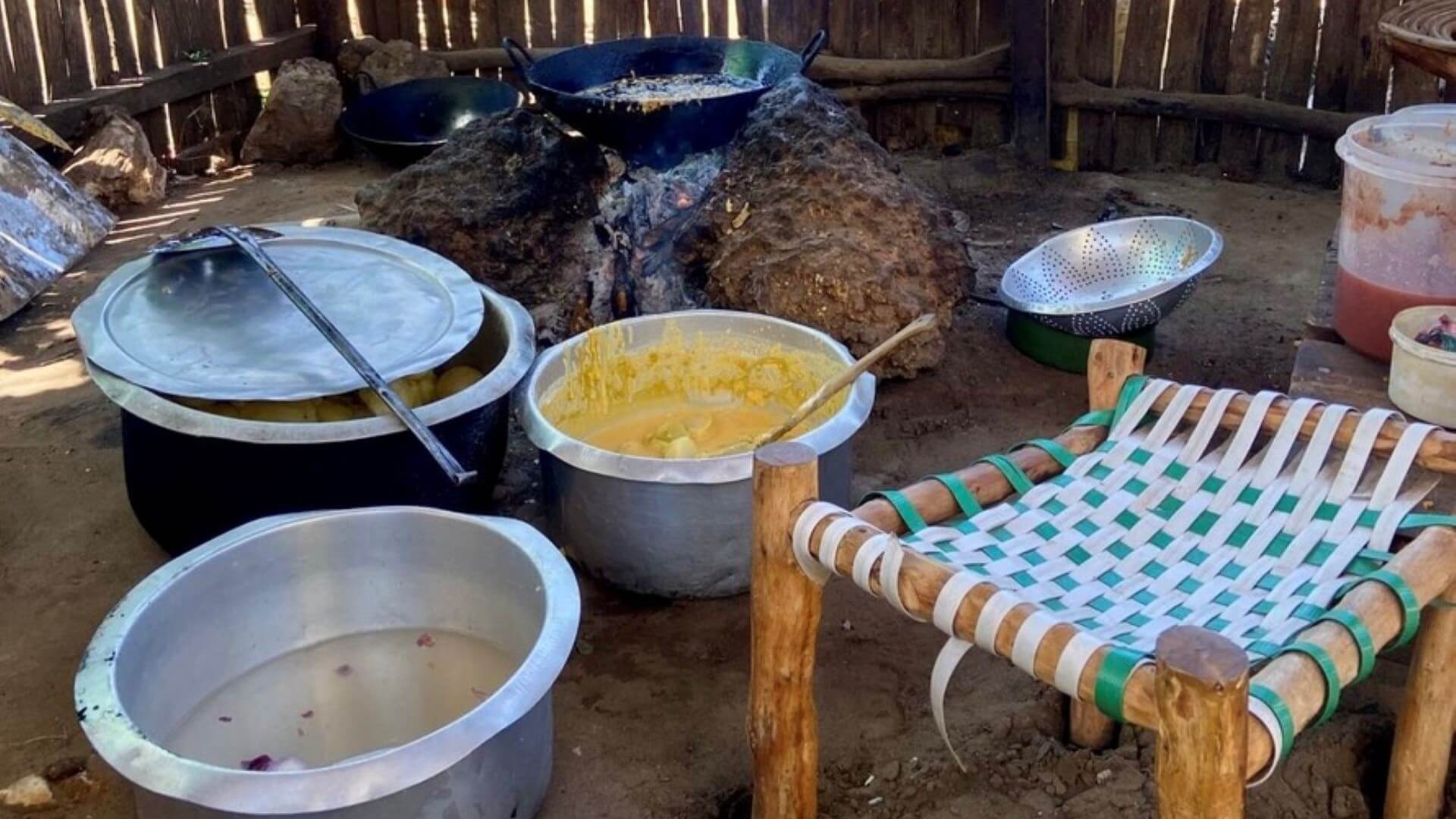 Various metal saucepans with food in, set on floor in front of an open charcoal fired stove with a black pan on top, cooking food, with a small stool also in the foreground