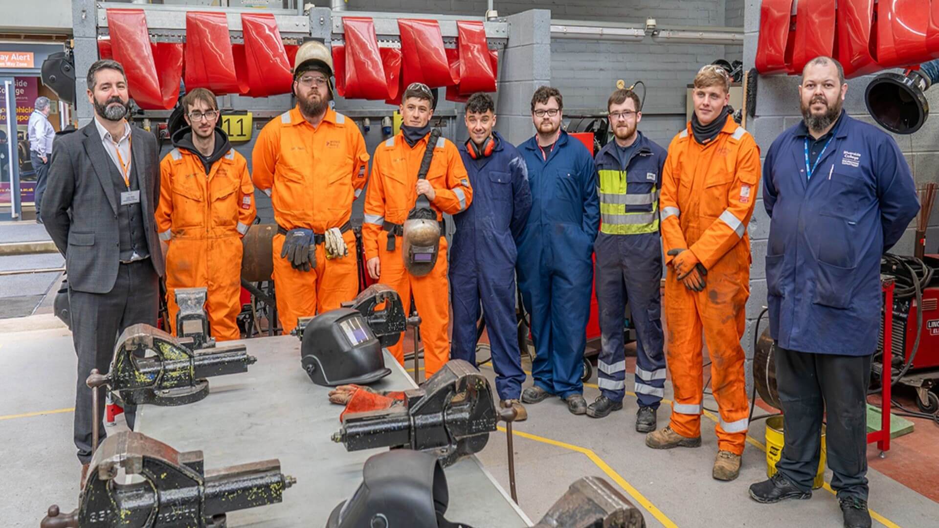 Row of nine men stood side by side, some in orange or blue safety boiler suits and overalls, and one in a business suit, surrounded by various equipment