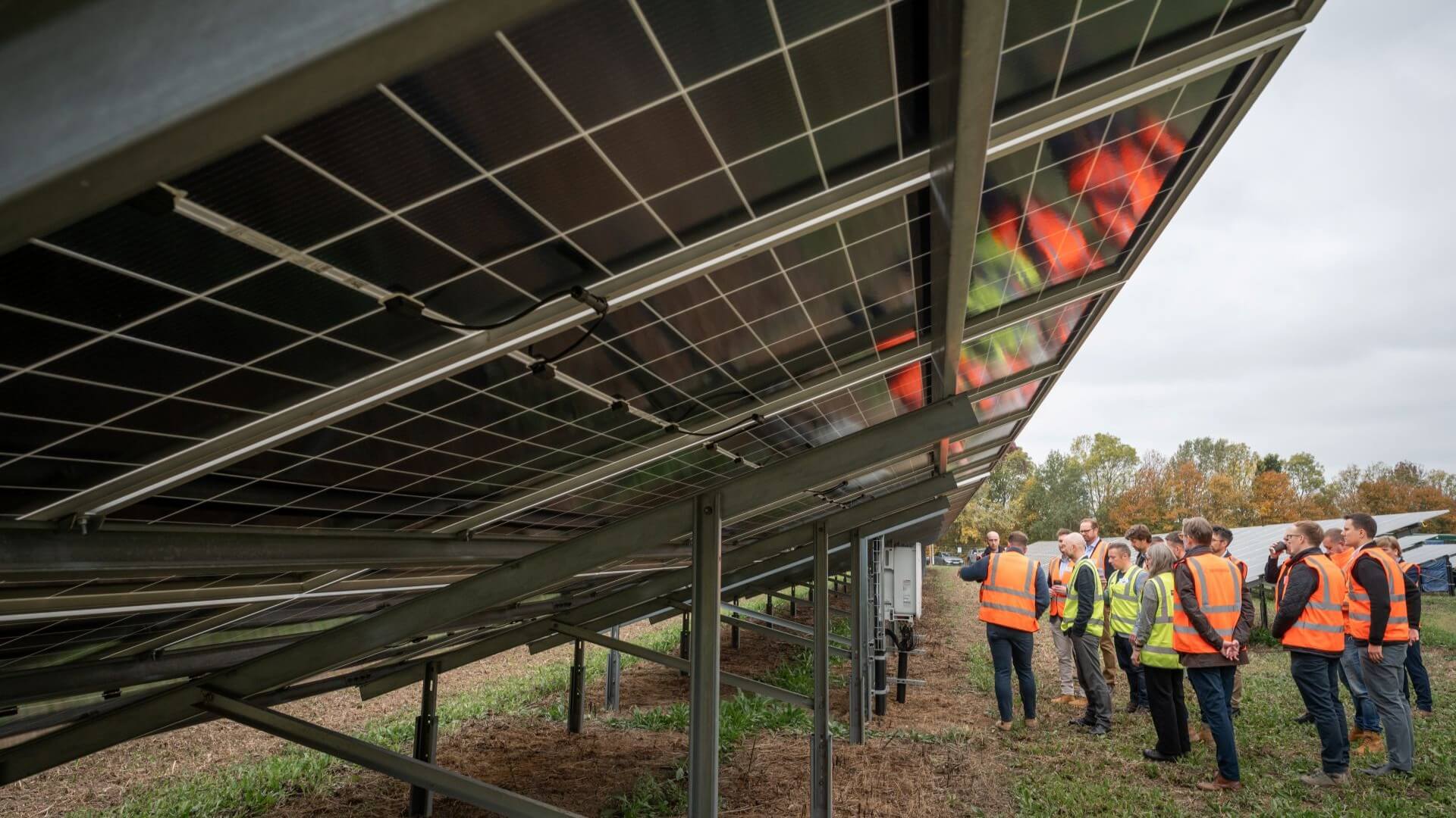 View along the underside of a row of solar panels, with a group of people in high-vis jackets standing close by and looking at the panels