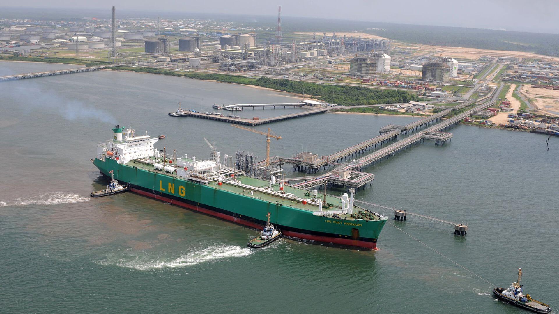 Aerial view of LNG tanker berthed at onshore terminal, with small tug boats to side
