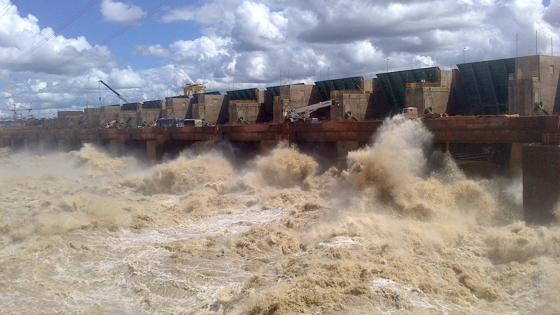 View along base of a dam wall, with raging brown and white water below and blue skies with while clouds above