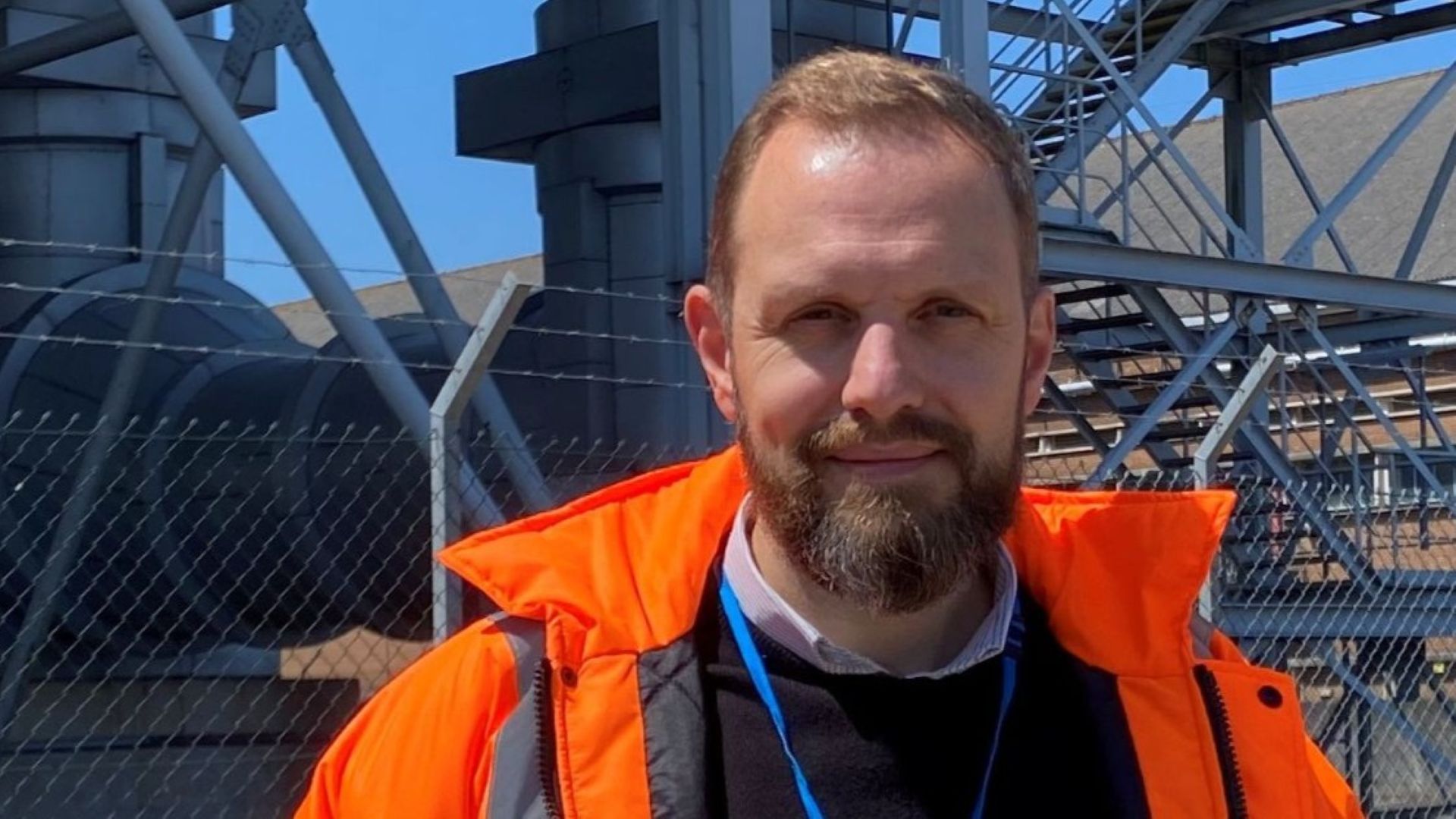Head and shoulders photo of John Burnett in orange high-viz jacket, with write mesh fence and close up electricity pylon behind.