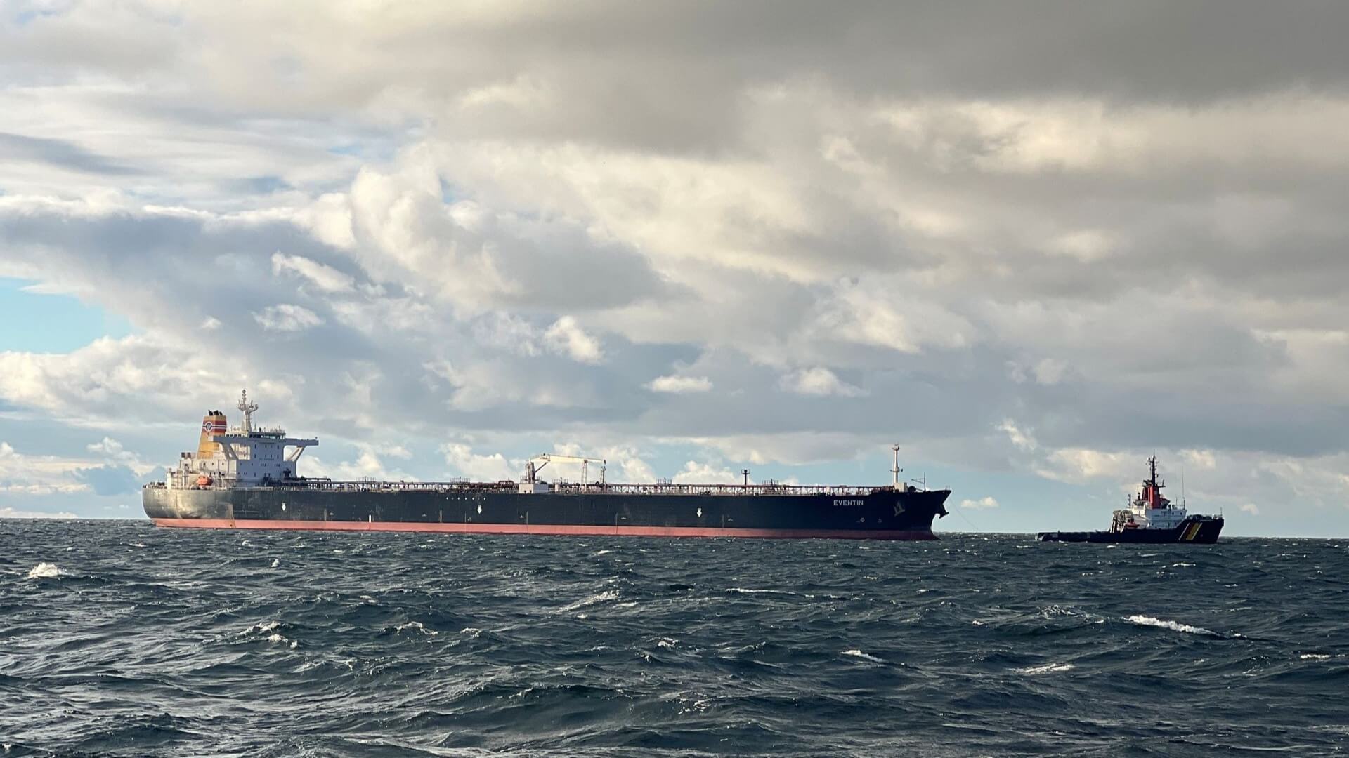 Distant view of large oil tankers with a small vessel towing from the front, in a grey sea with grey clouds above