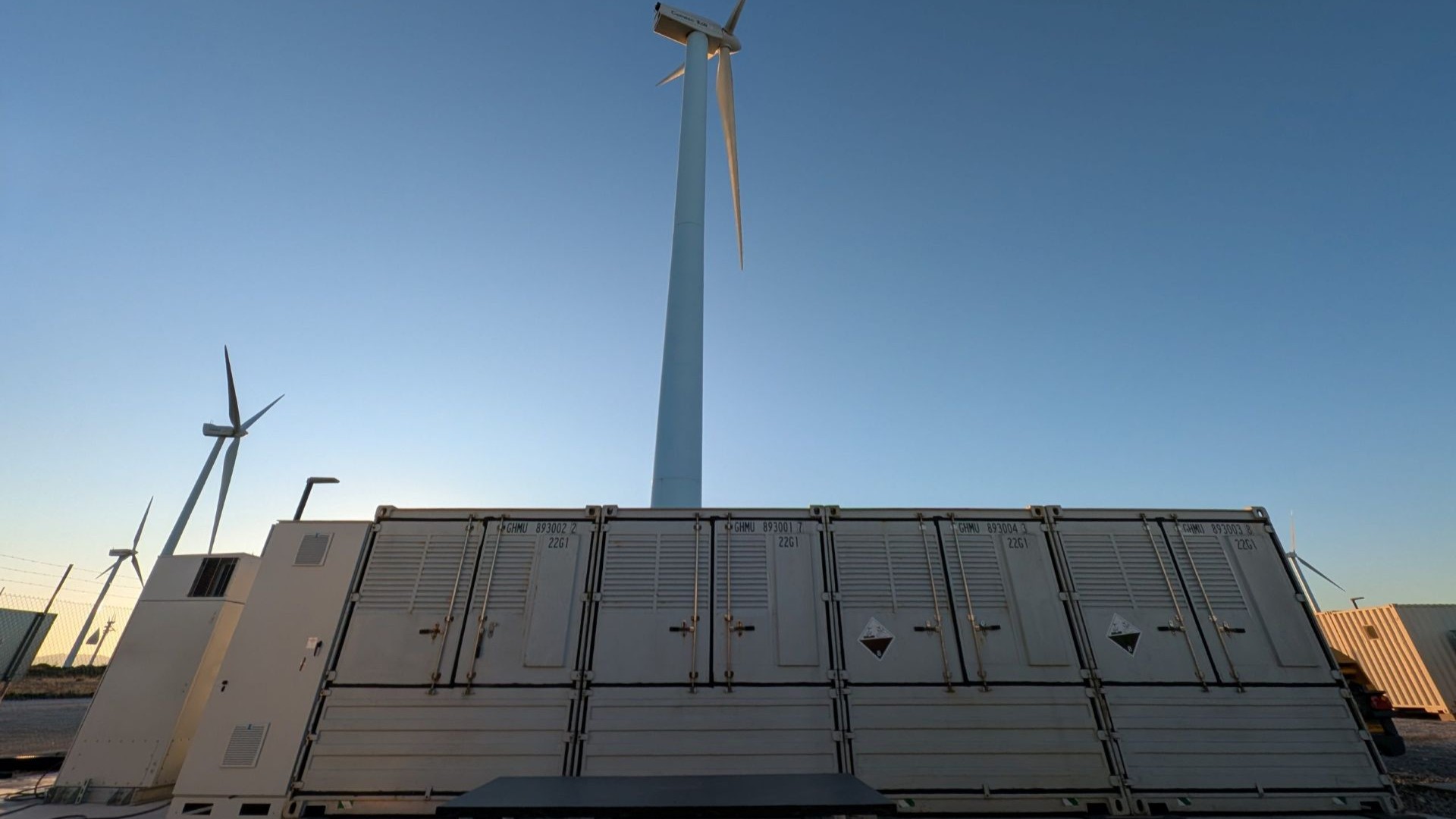 Photo taken from ground at base of a row of large rectangular shaped metal boxes housing batteries, looking up at a wind turbine standing behind and others in the background to the left