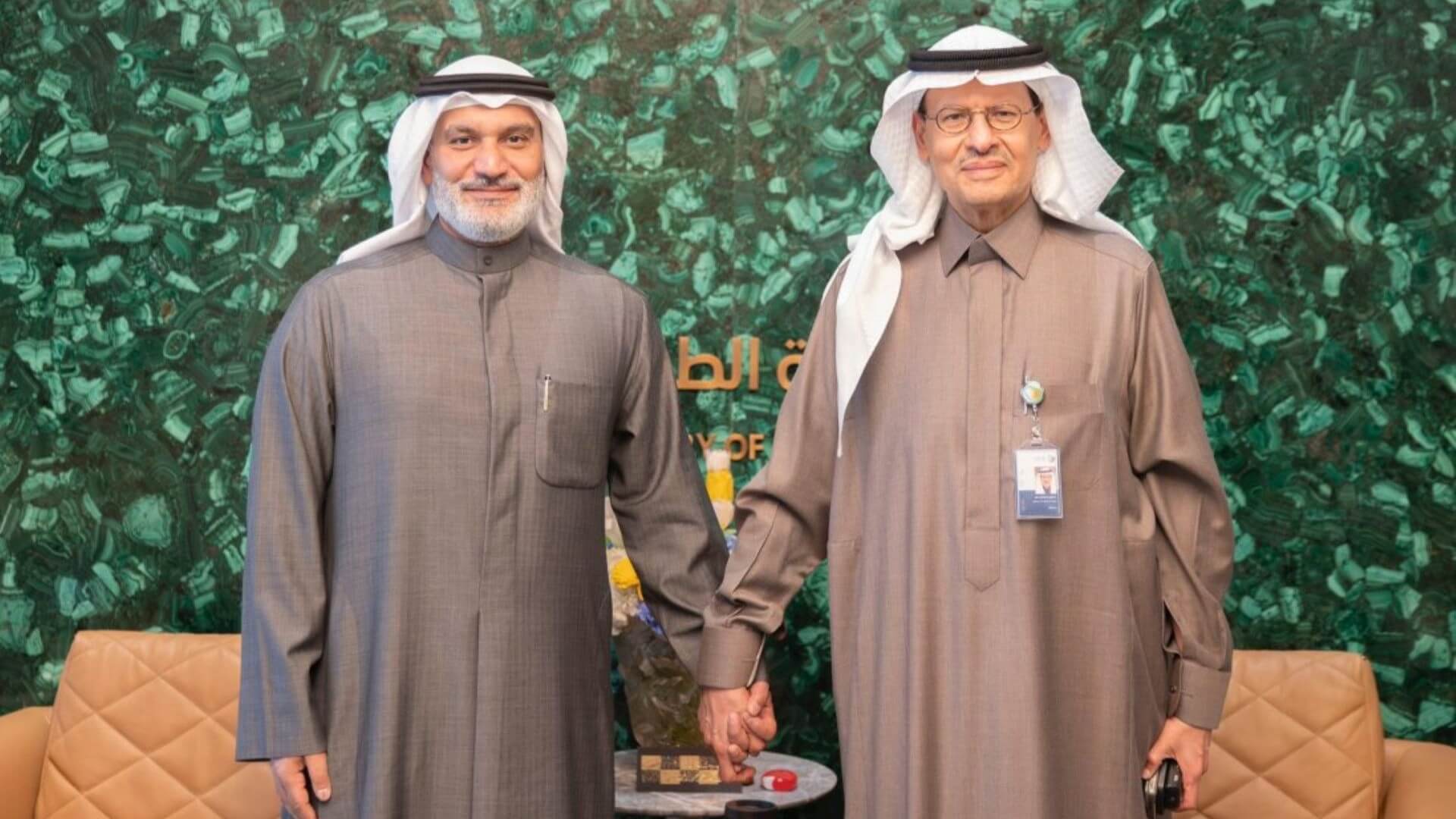 Photo of two men in traditional Arabic clothing, standing side by side in front of a backdrop of green foliage