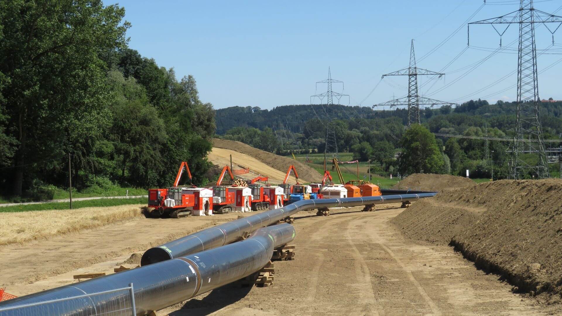 View along a line of silver pipe sitting on wooden blocks on an earth track, awaiting installation, with a row of caterpillar tracked vehicles with lifting arms in the distance and line of electricity pylons and transmission wires to the right