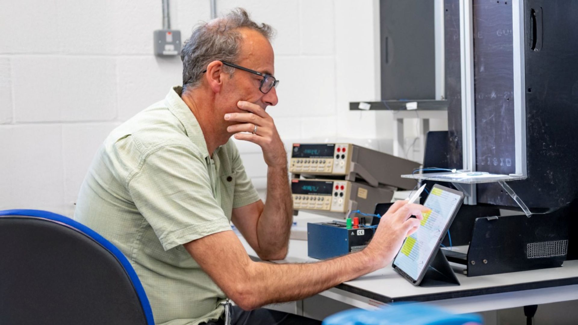 Side on head and shoulder photo of Professor Patrick James sat a desk with left hand cupping chin and elbow resting on desk, while his right hand is keying something in to an angled touch screen; various lab equipment and wall and window in background