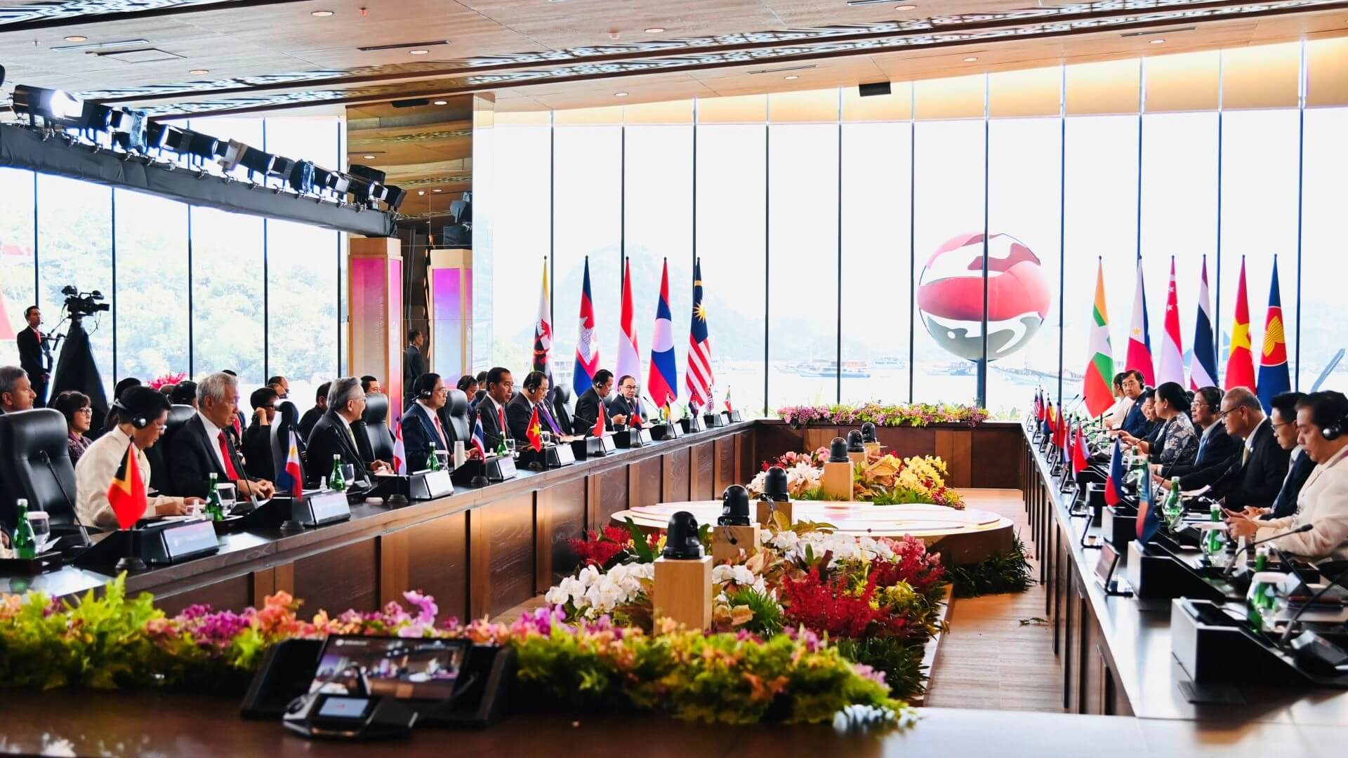 Rows of people sat at tables set in rectangular formation, with windows in background and national flags