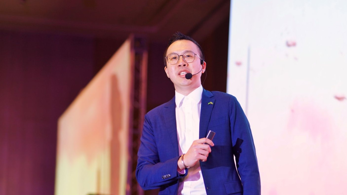 Dr Lim Eu Shawn holding powerpoint clicker and wearing microphone headset, standing on stage with screen to back left, talking to conference delegates