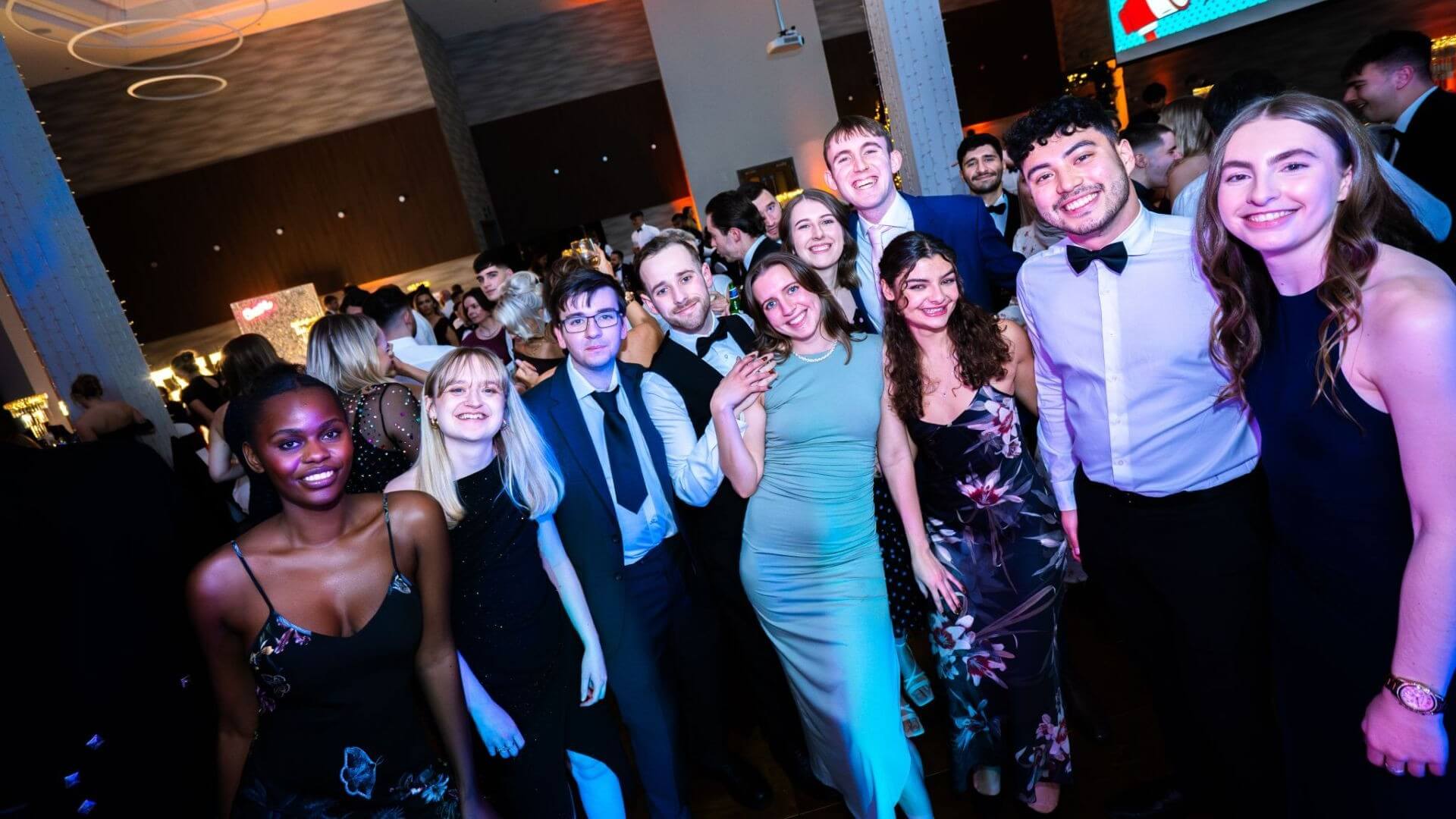 Group of young people in black tie and cocktail dresses smiling to camera, against background crowd of people socialising at dinner dance