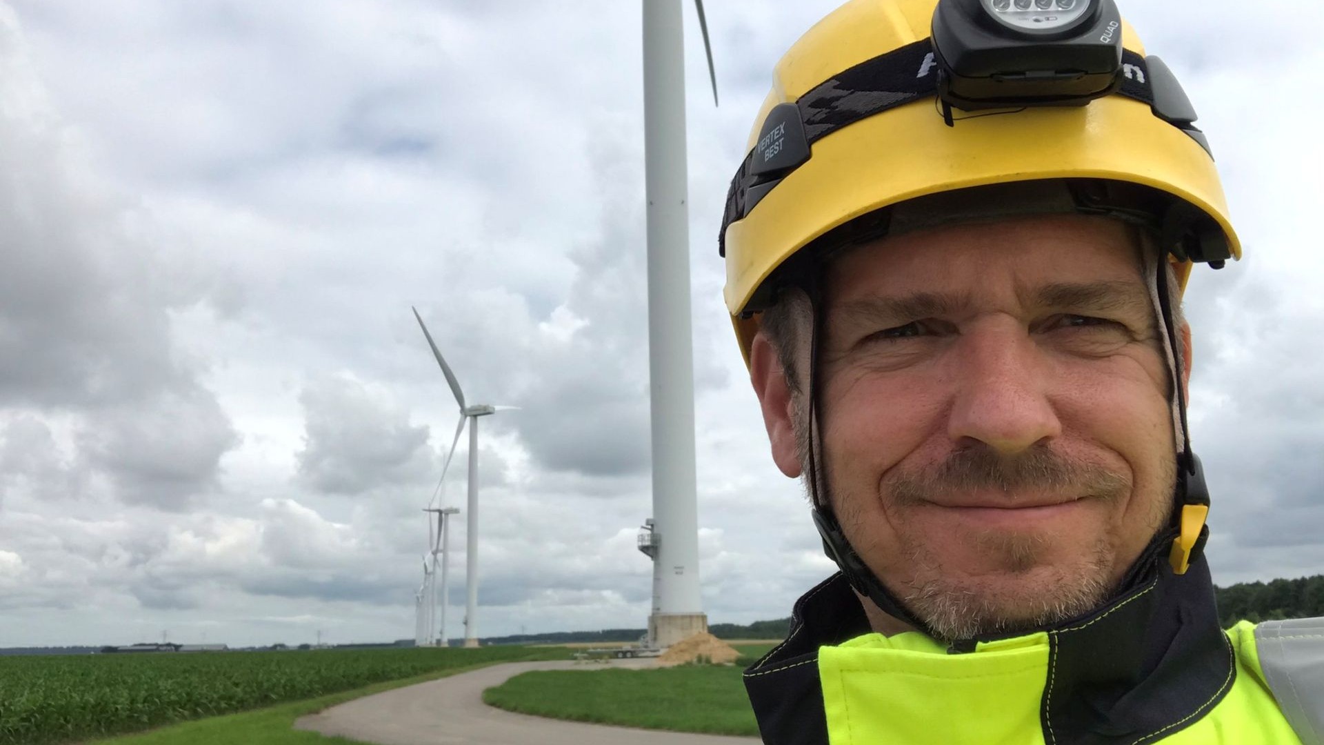 Close up head and shoulders photo of Darren Taylor in right foreground of picture, with a row of onshore wind turbines behind and extending into the distance