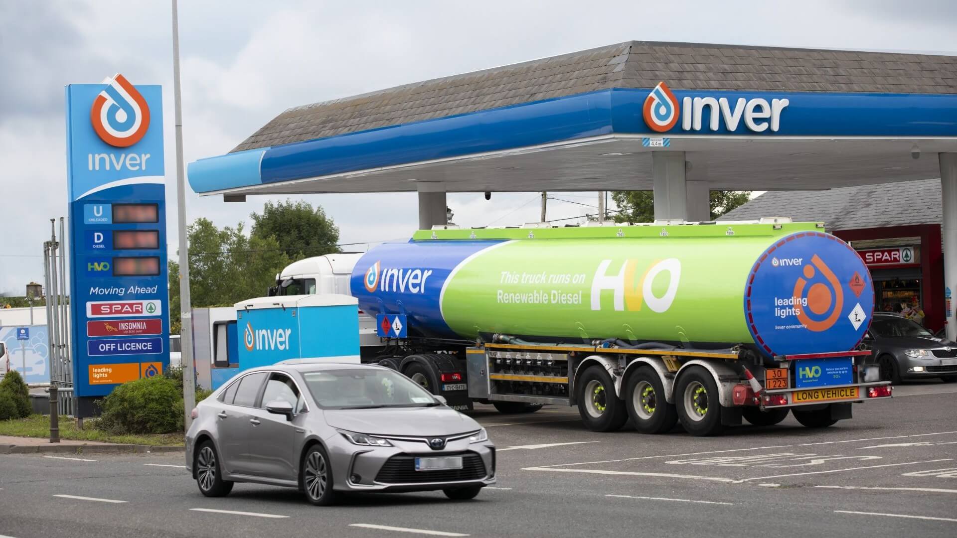 View of petrol station with pricing board to left and a fuel tanker driving onto the forecourt, with a car passing on the main road alongside the service station