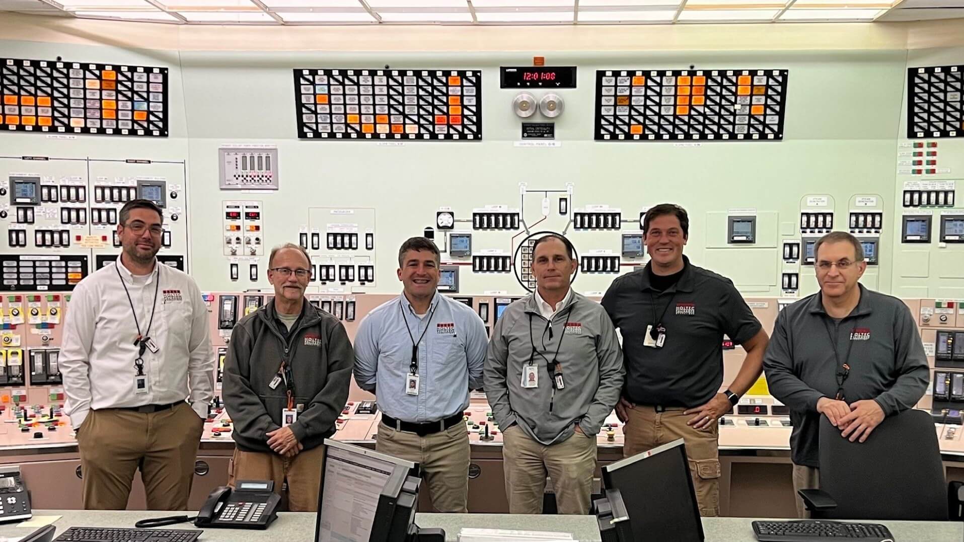 Row of six men standing side by side, smiling, in a control room with banks of switches and dials behind and long counter with computer monitors and keyboards in front