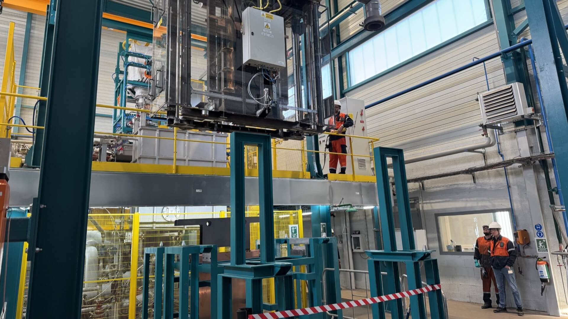 Inside view of industrial plant with metal work and railings and lifting device, with a few men standing to side in orange high-viz boiler suits and safety helmets