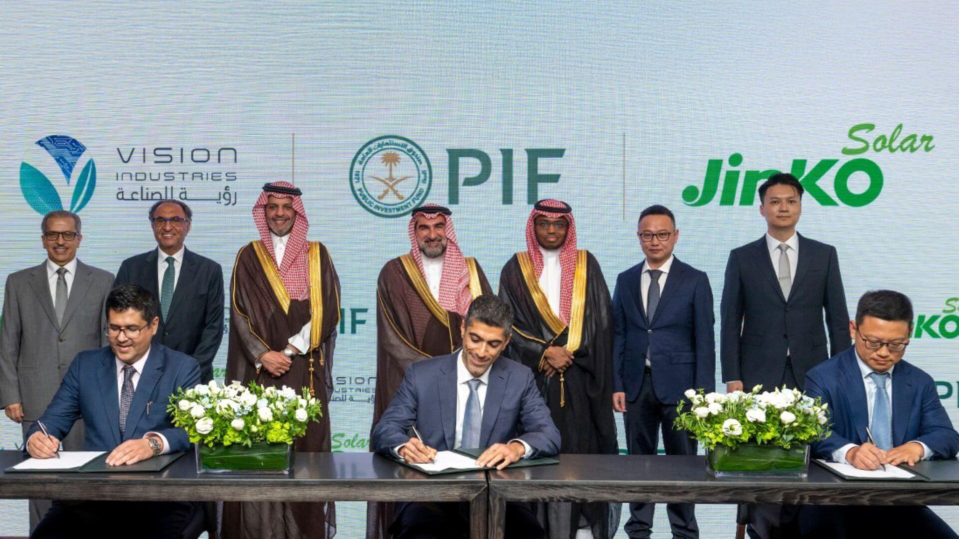 Three men in suits, sat at long table and signing documents, with row of seven company executives standing behind them, with a backboard showing company logos