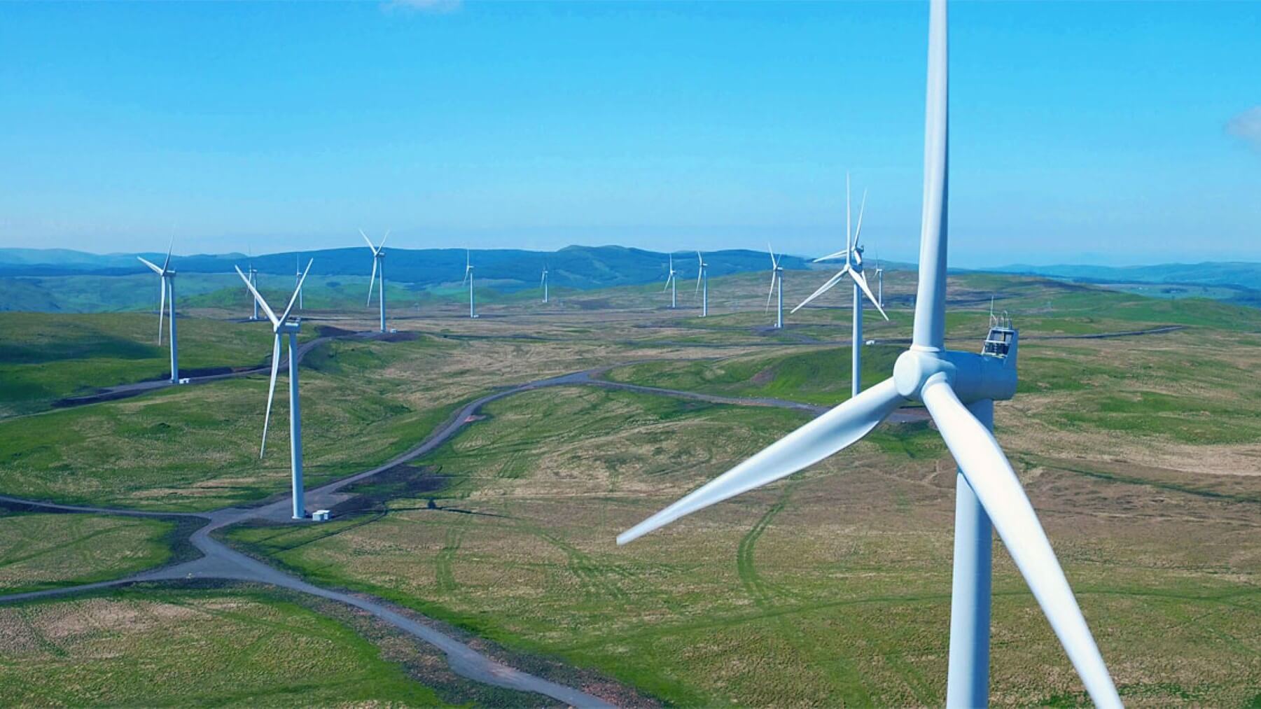 Aerial view over an onshore wind farm, with one turbine in the foreground and other turbines in the distance, set against a blue sky