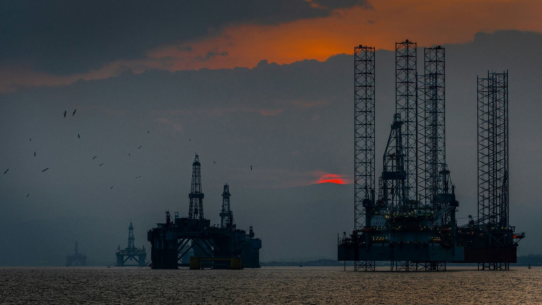 View across calm flat water with a number of oil drilling rigs parked fairly close together and silhouetted against a dark sunset sky