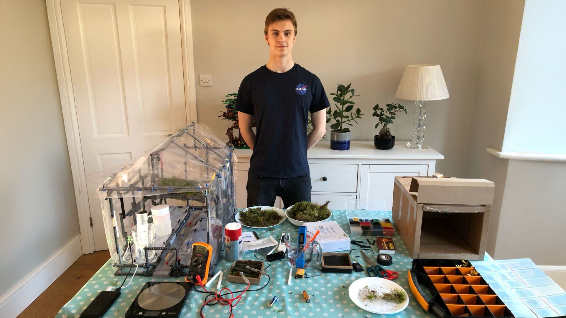 Oliver Linehan standing in front of table in front room of home, with component parts of his invention spread across the table top