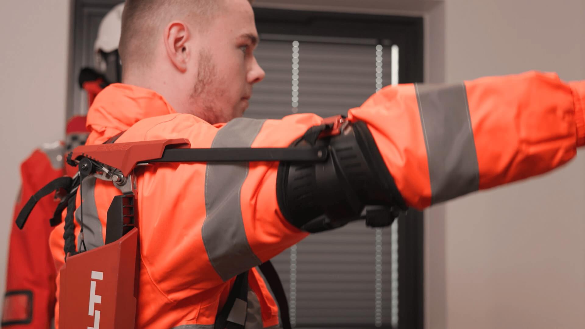 Close up of man in orange boiler suit, wearing an exoskeleton brace on back and attached to elbow of raised arm
