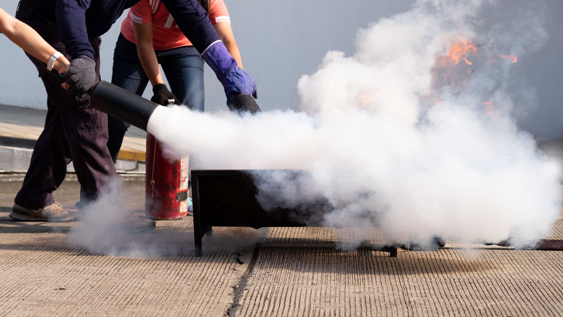 Close up of two people in safety gear tackling a fire contained in a small metal container with a fire extinguisher, with plumes of CO2 gas billowing from extinguisher nozzle