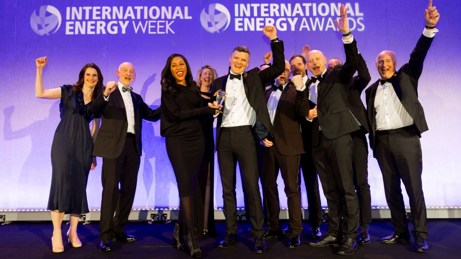 Group of men and women, in smart evening attire, standing on stage with smile and some with fists raised in the air in celebration