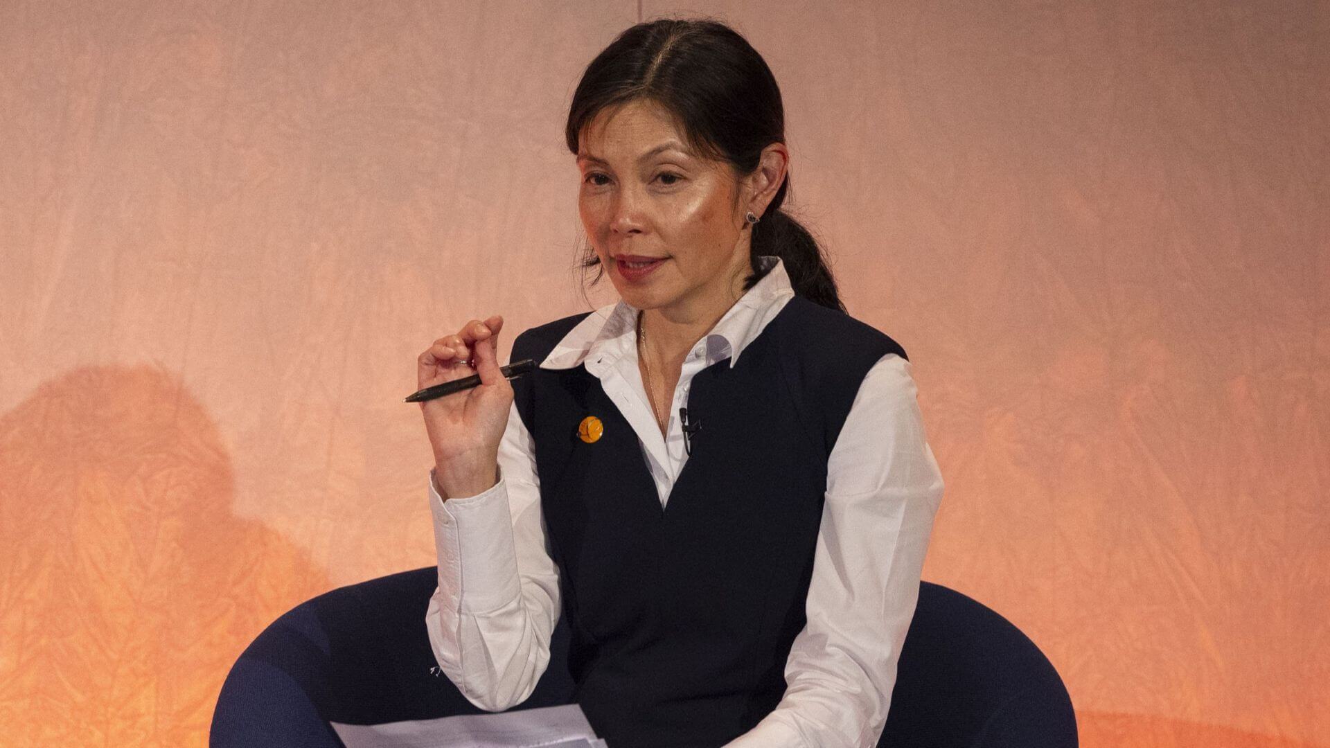 Head and shoulders photo of sitting in a chair on stage, right hand raised while talking over a point with conference delegates