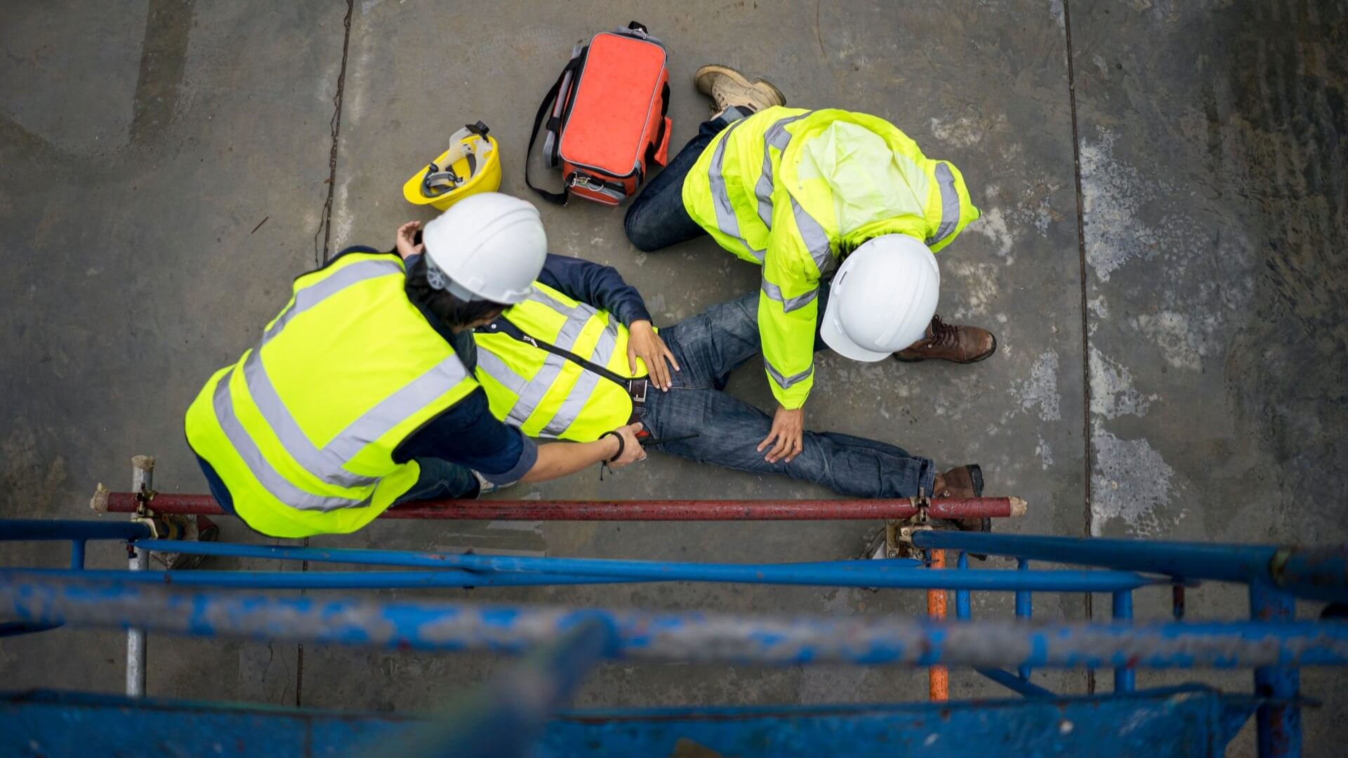 Bird's eye view of two emergency responders in high-viz yellow jackets and white hard hats attending a person lying on a concrete floor outside, having fallen from a scaffolding tower