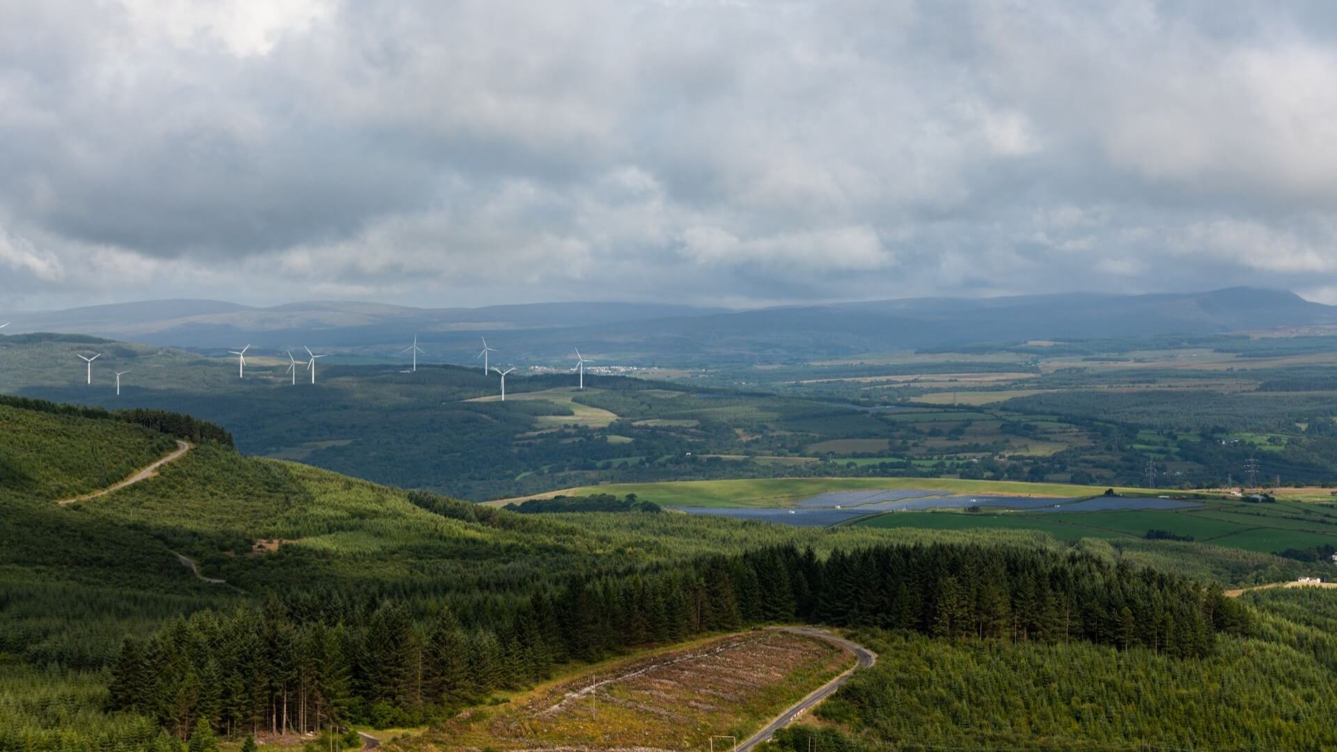 Aerial view of wind farm with lots of turbines, and solar farm with row upon row of solar panels, in the distance set amid green fields and countryside