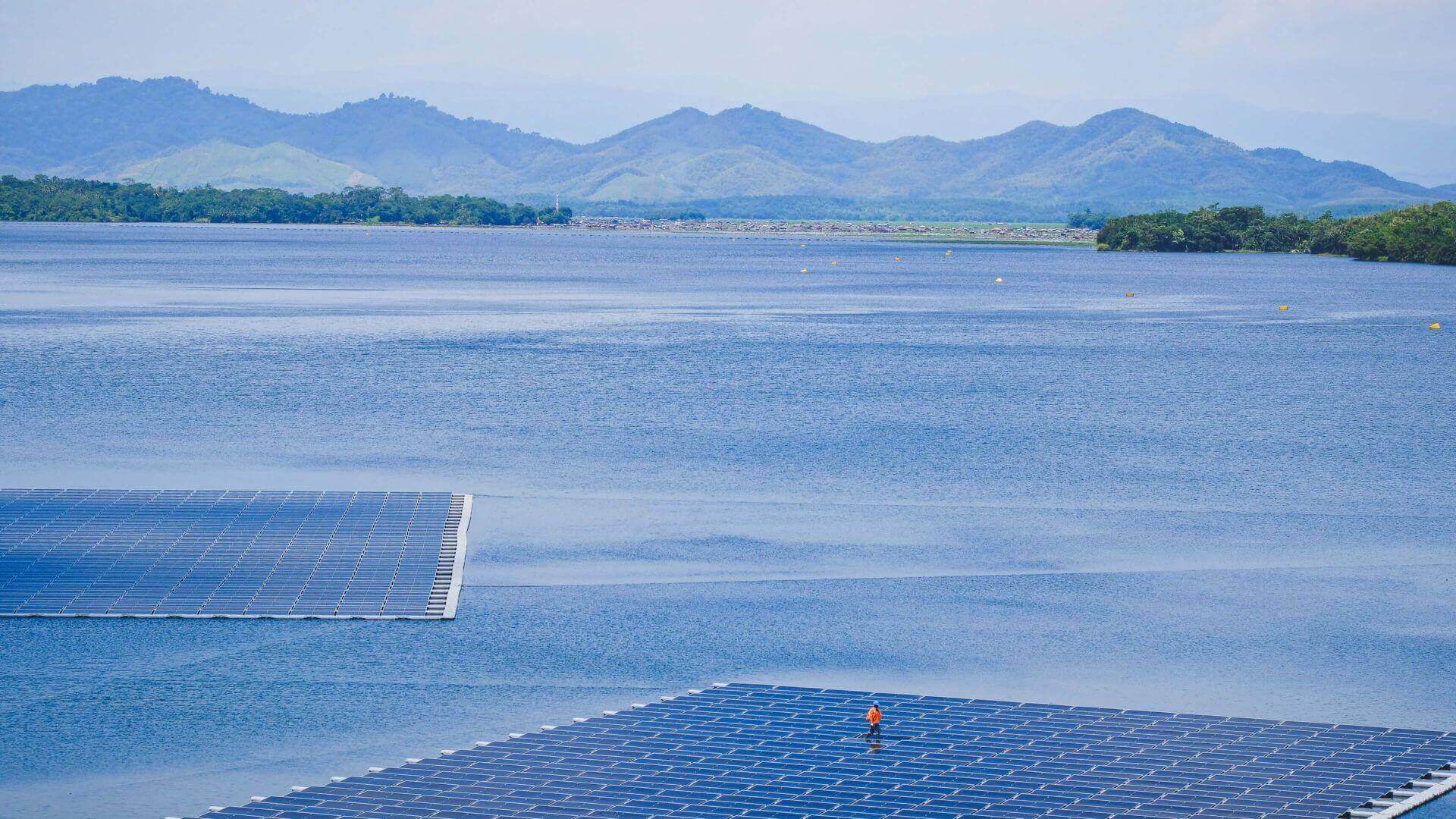 Aerial view over floating solar farm in foreground, with an operator standing in the middle of the farm; rest of lake and mountains in the distance
