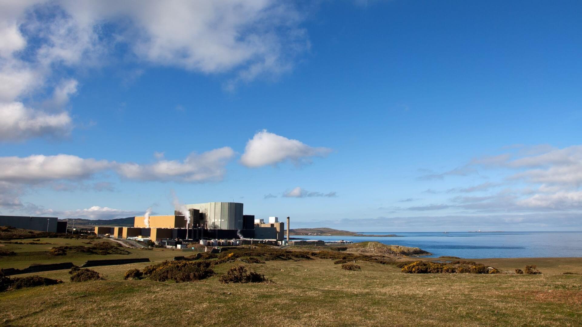 Distant view of large, blocky nuclear plant buildings set on coastline, with green land to foreground and sea in background, blue skies and white fluffy clouds above