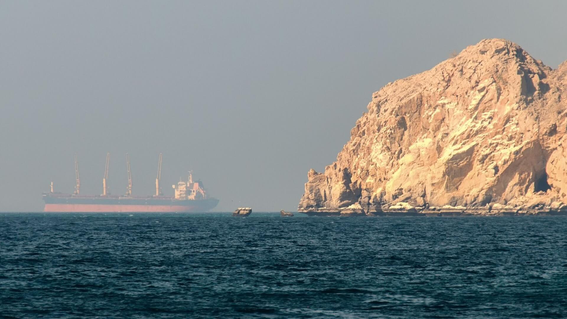 Distant view of a large ship in calm sea to left of picture, with cliff face to right of picture
