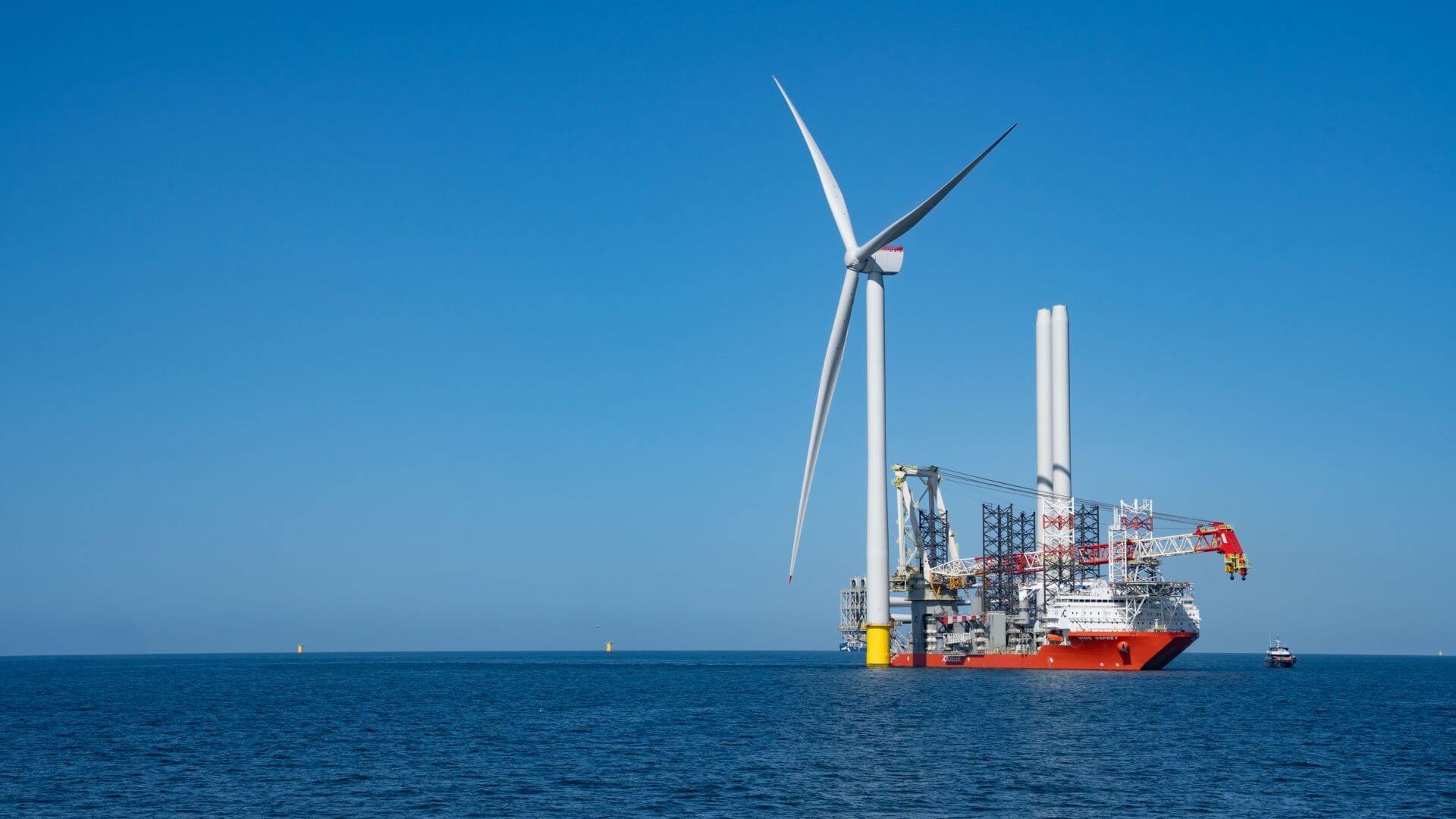 Installation vessel alongside three-bladed turbine, in calm sea and set against blue sky