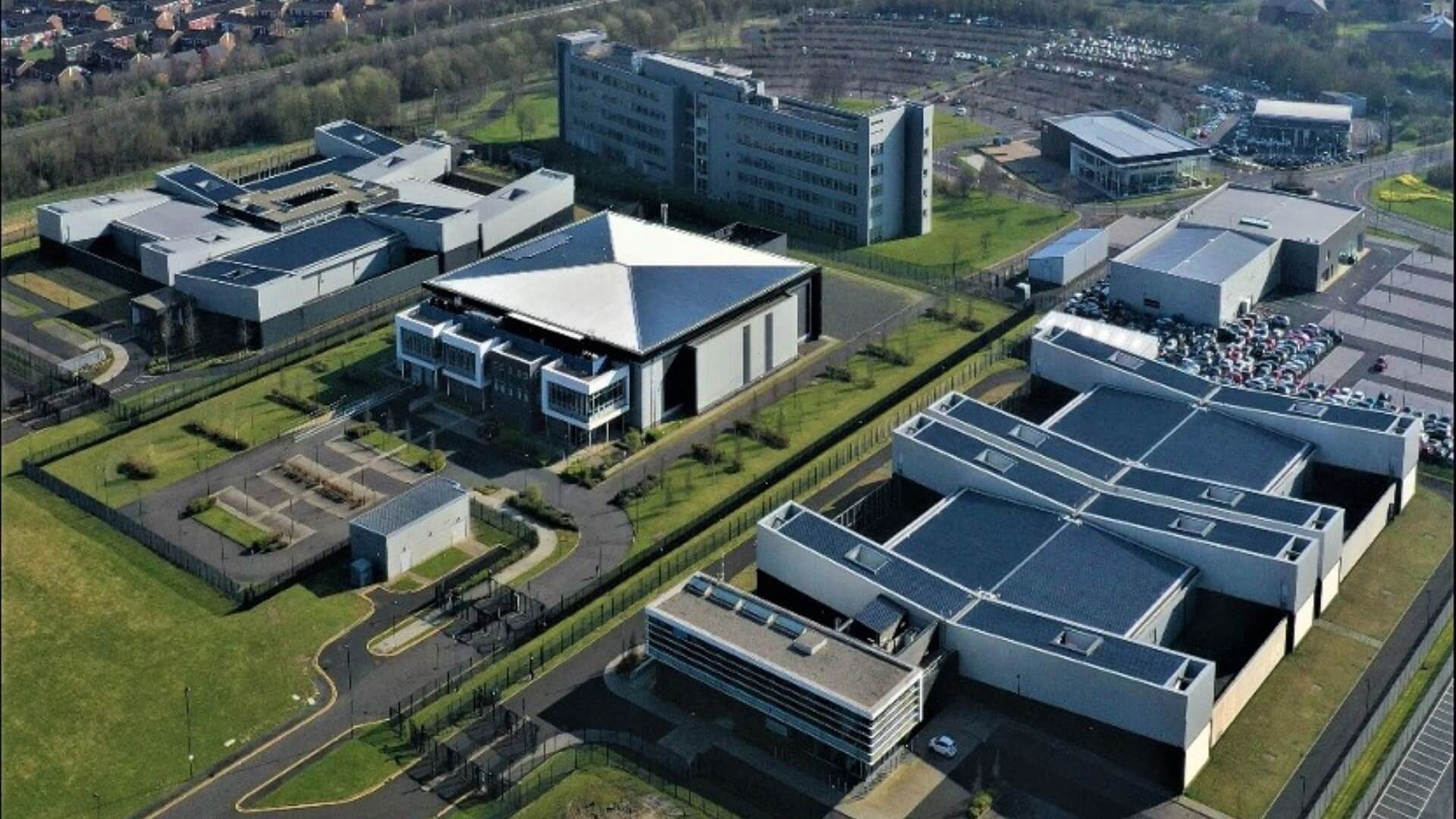Ariel view over the Stellium data site, showing series of rectangular buildings with angular sloping roofs, surrounded by green space and trees, and car parks