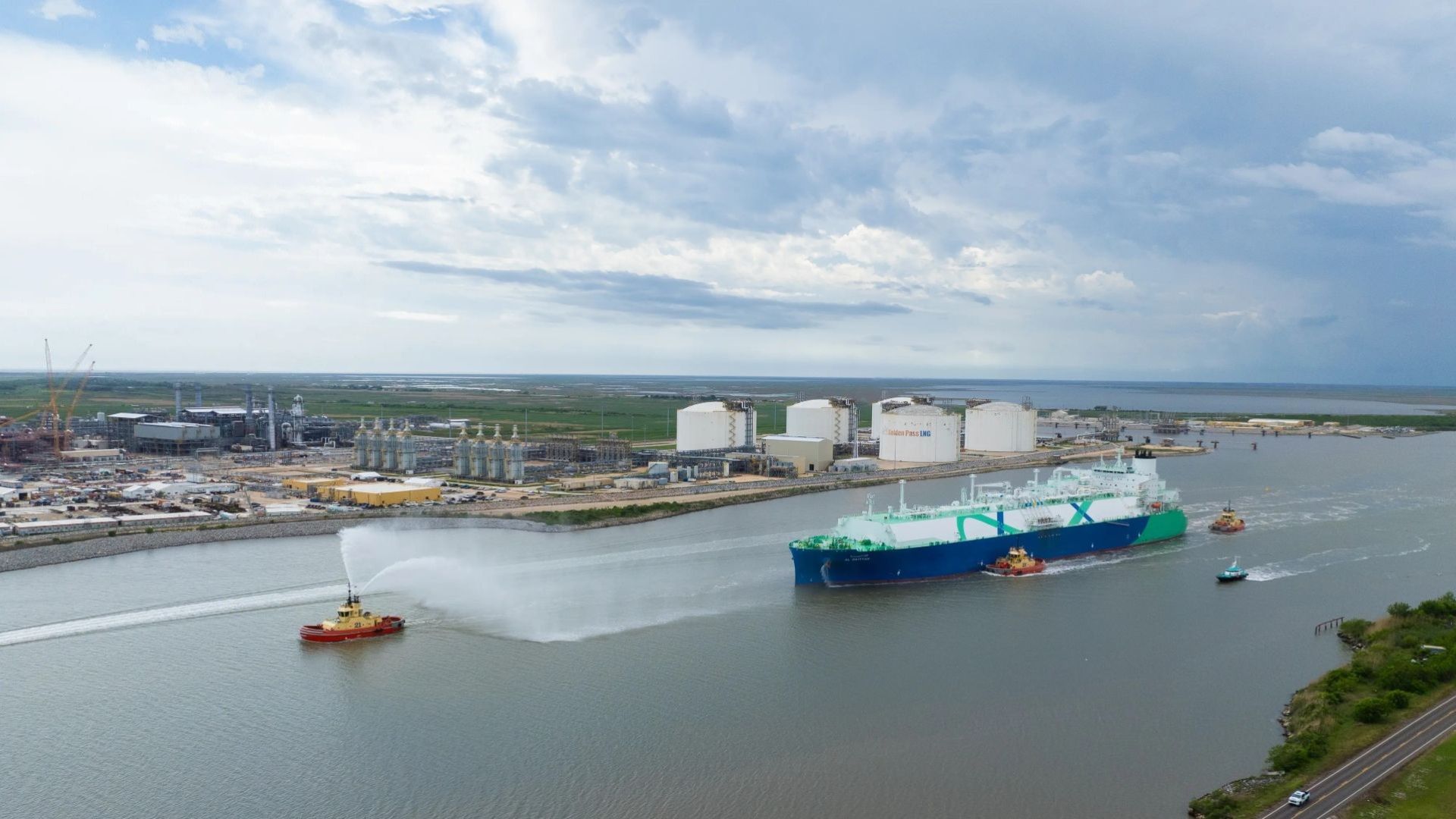 Aerial view of gas tanker accompanied by pilot boat and a few smaller vessels at terminal, with LNG storage tanks and terminal facilities in the background