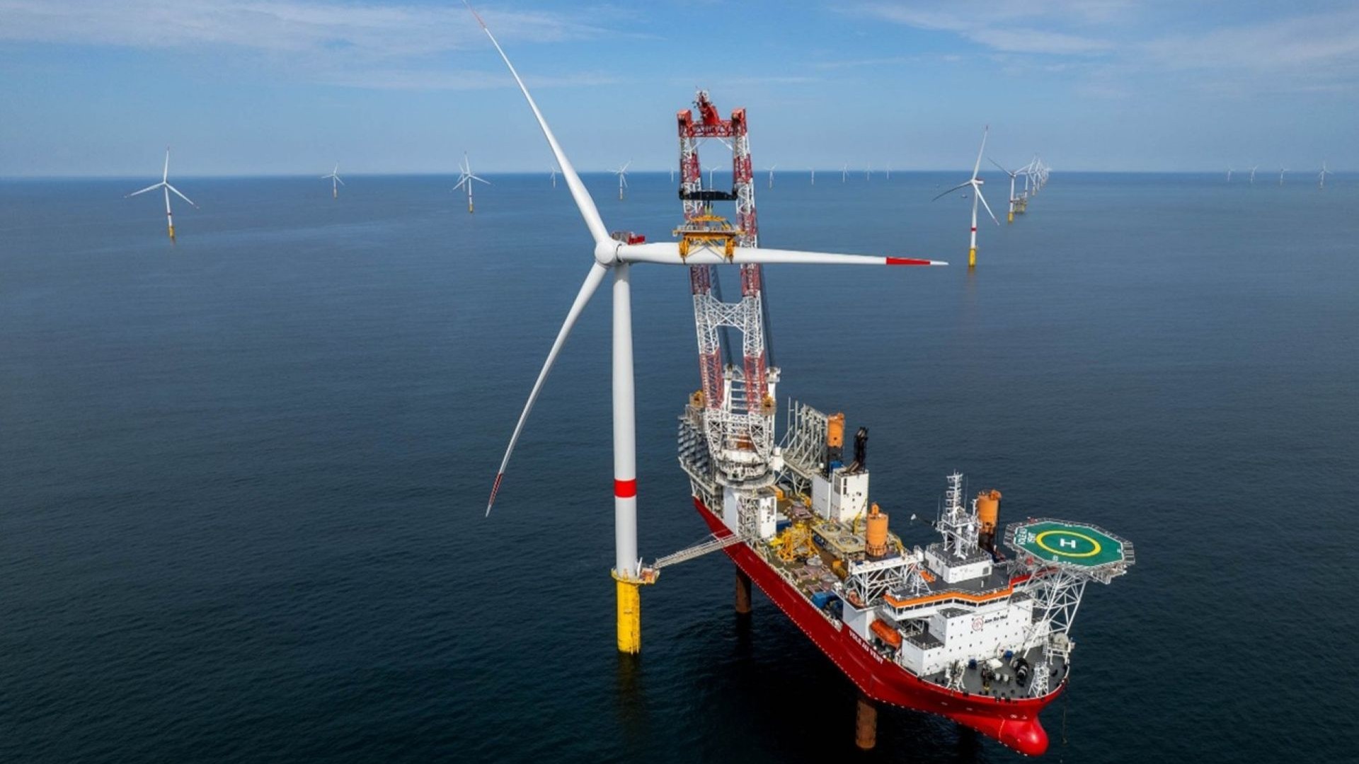 Aerial view of third blade being installed on an offshore wind turbine, by a vessel in calm sea, with other offshore wind turbines in the distance