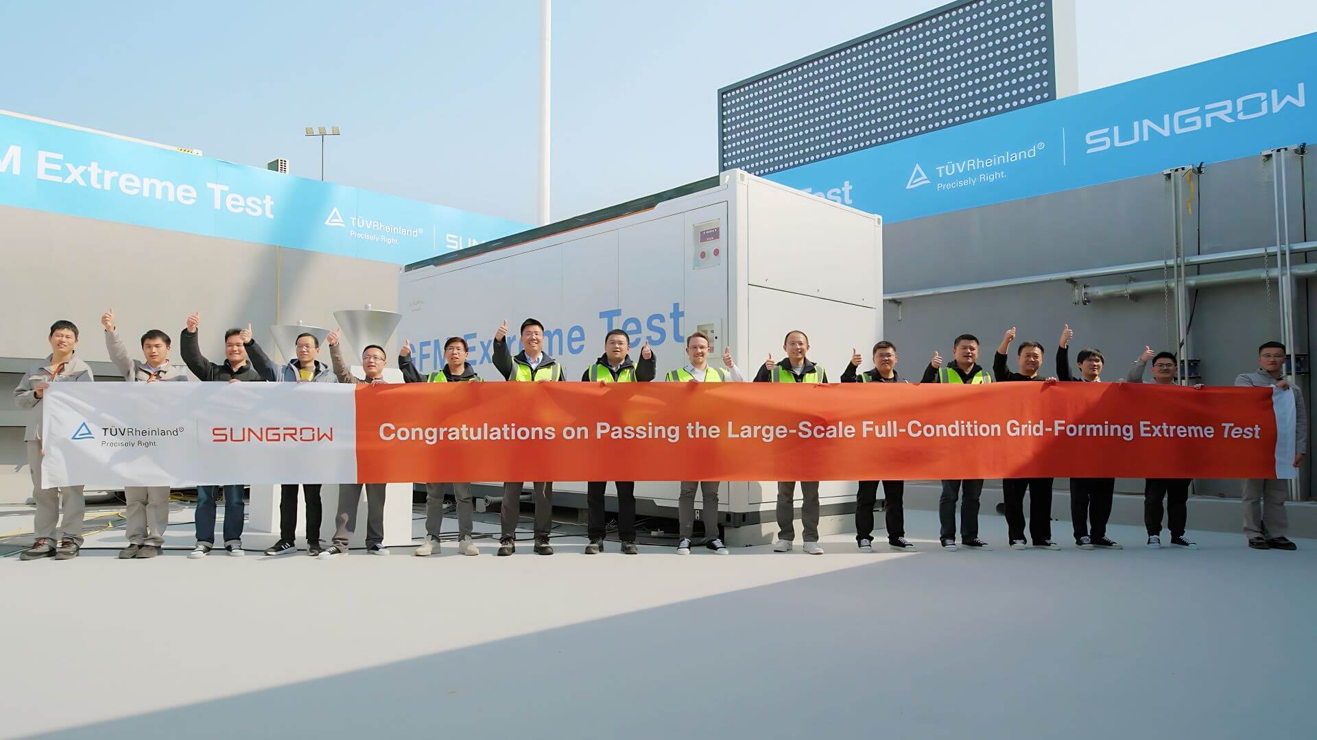 Sixteen people standing in a row, holding a very long banner with two company logos and a congratulatory message regarding successful test results; large container and office building in background