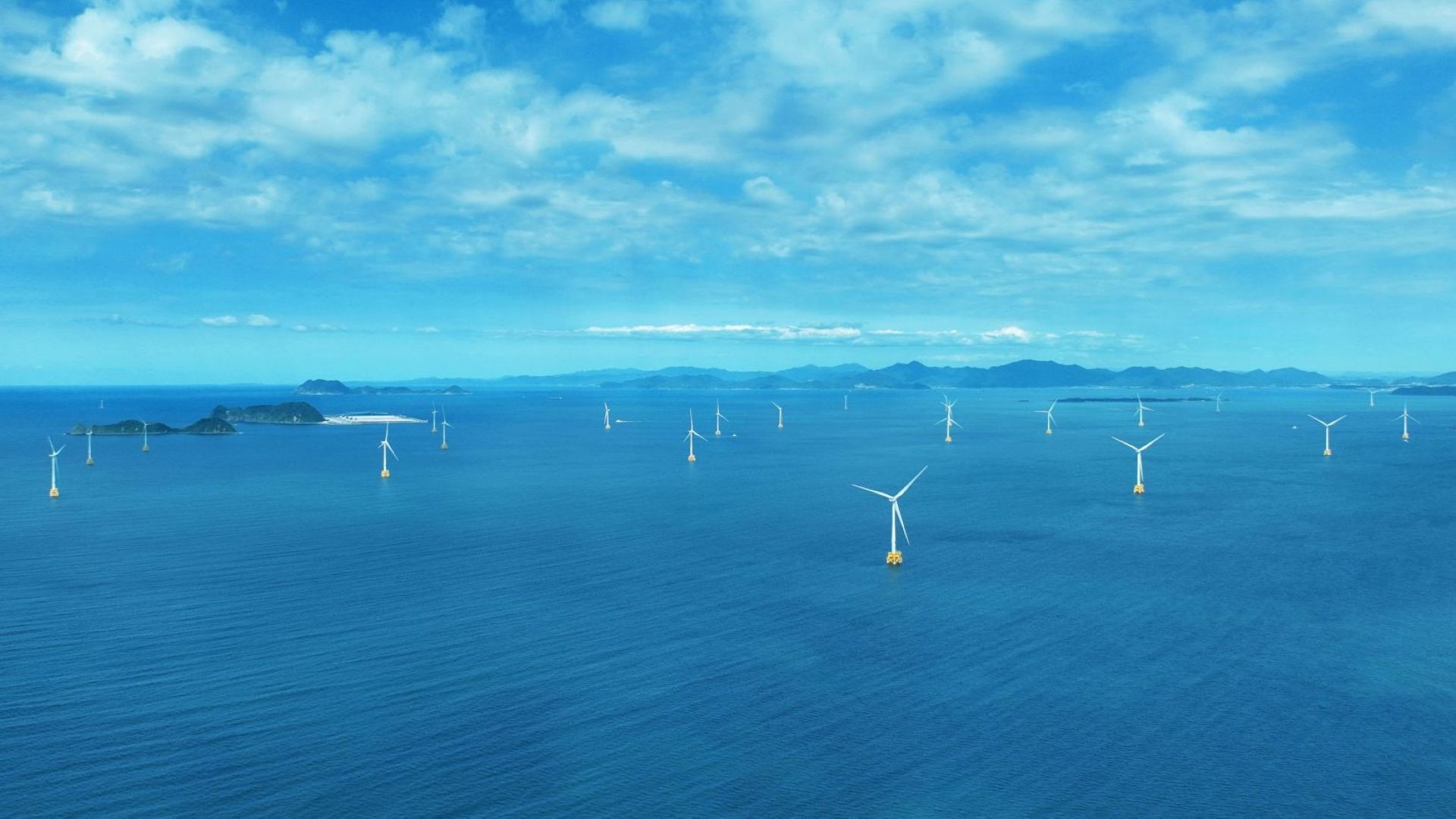 Aerial view over offshore wind farm, showing rows of wind turbines in calm blue set, set against blue ski with white fluffy clouds