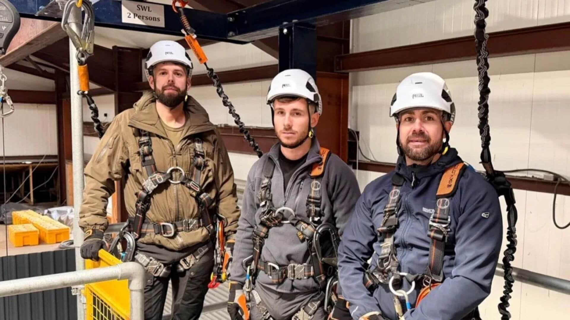 Three men in a row, one standing, two leaning against a metal handrail, all wearing hard hats and safety gear