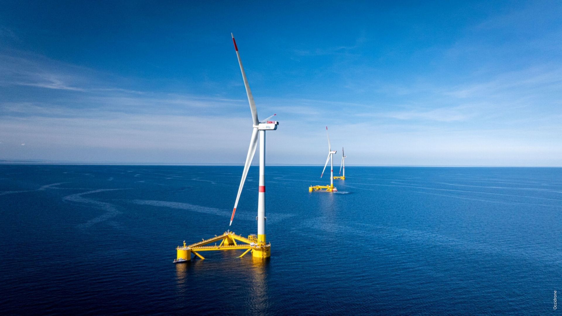 View of three floating offshore wind turbines in a row, extending into distance, in calm blue sea and set against blue sky