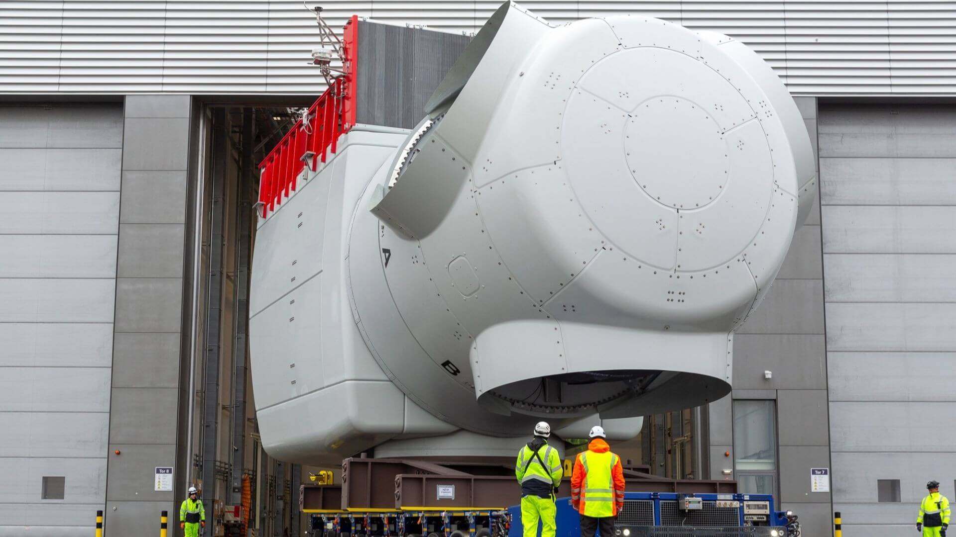 Large wind turbine nacelle being moved out of fabrication warehouse, with little room to spare on exit, and dwarfing a few people in high-vis gear and hard hats that are standing nearby