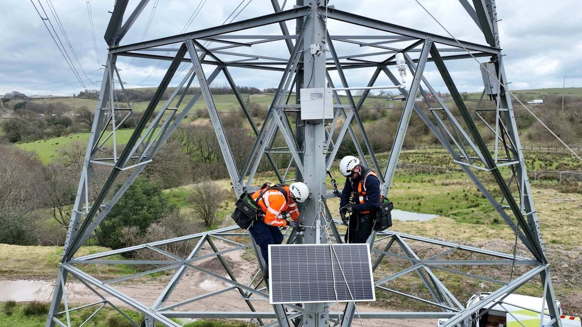 Two engineers in hard hats and safety equipment, half-way up the metalwork of an electricity pylon, installing a small rectangular box of equipment and solar panel to power it