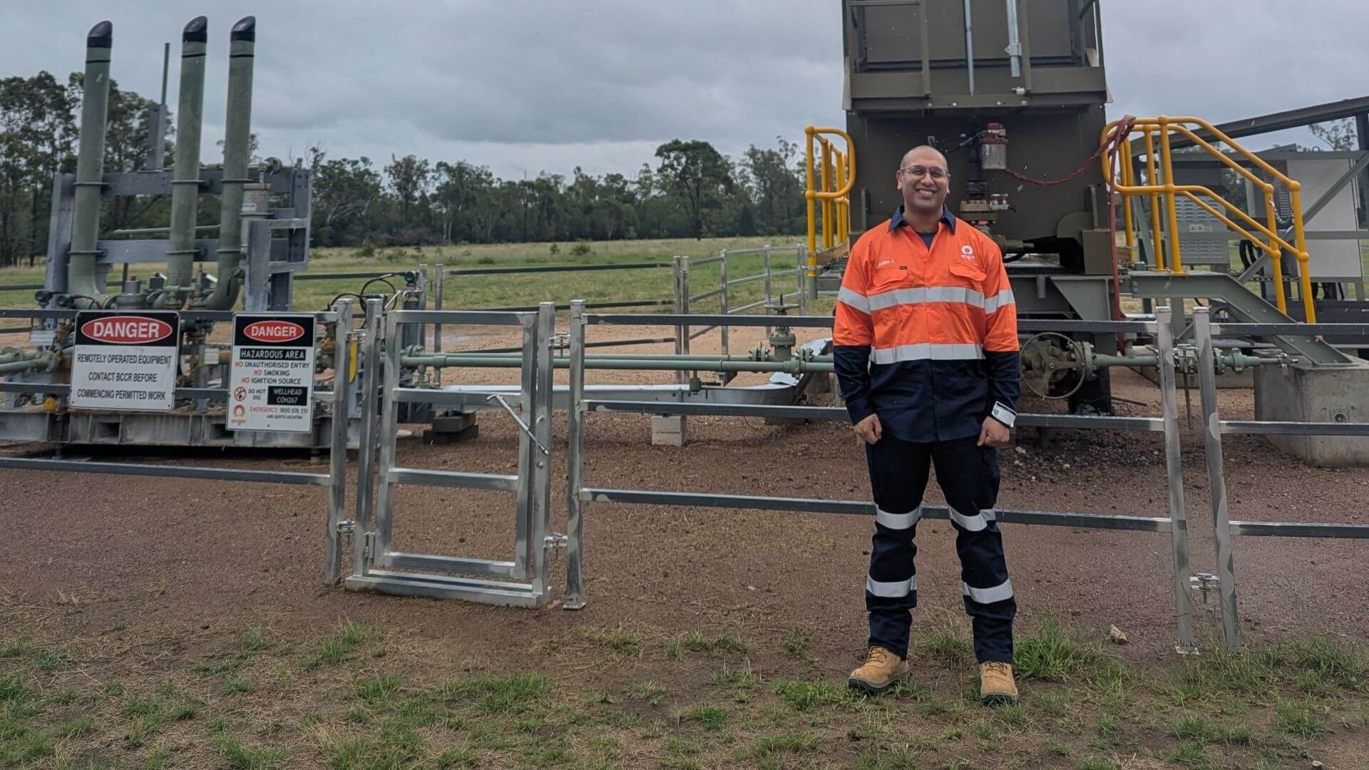 Zubin Jehangir in high-vis jacket and work clothing, standing outside in front of metal fence surrounding industrial equipment