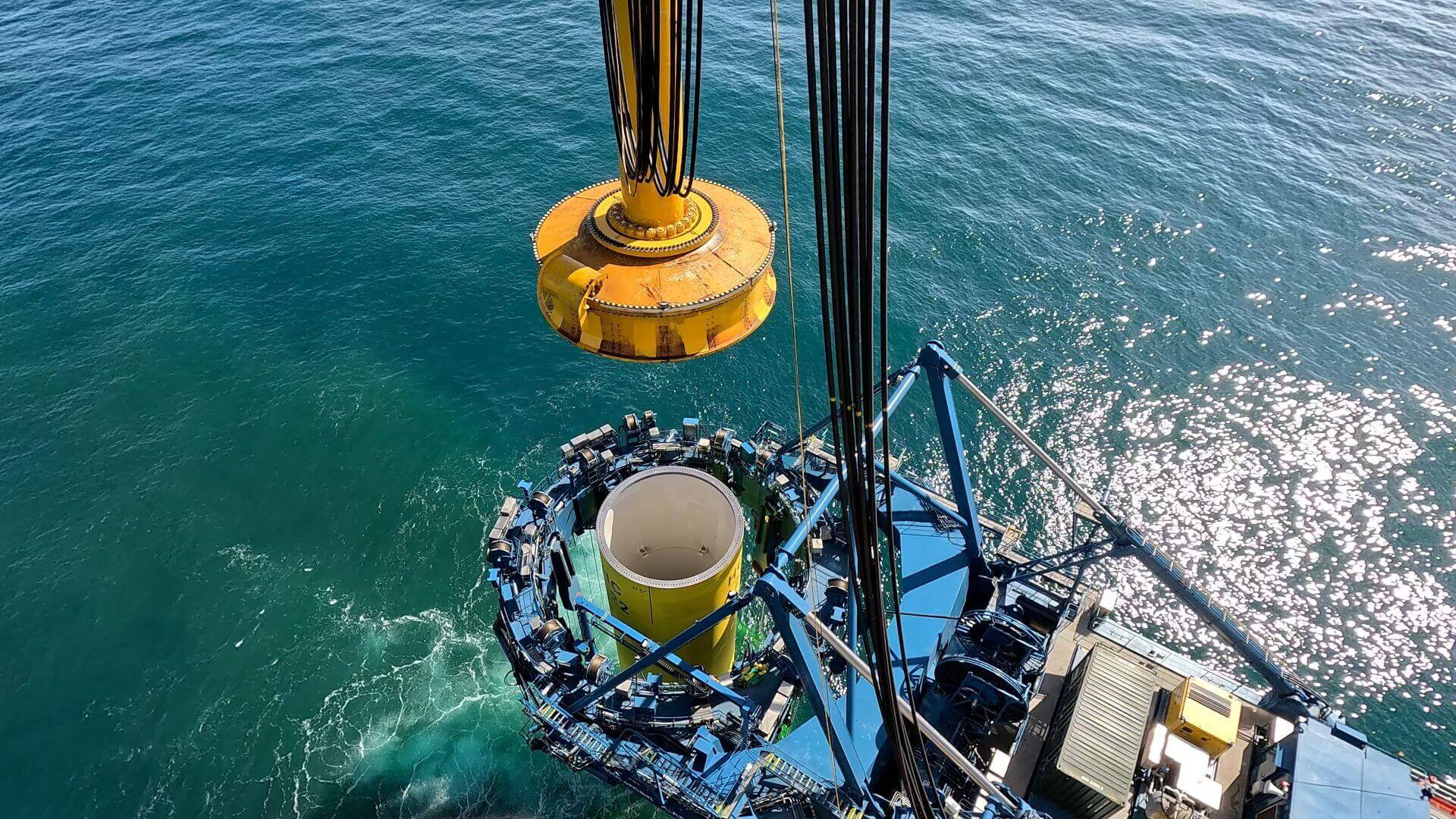 Close up aerial view of monopile tube being held by a giant claw clamp with a circular 'cap' being placed on top to push the foundation into the seabed