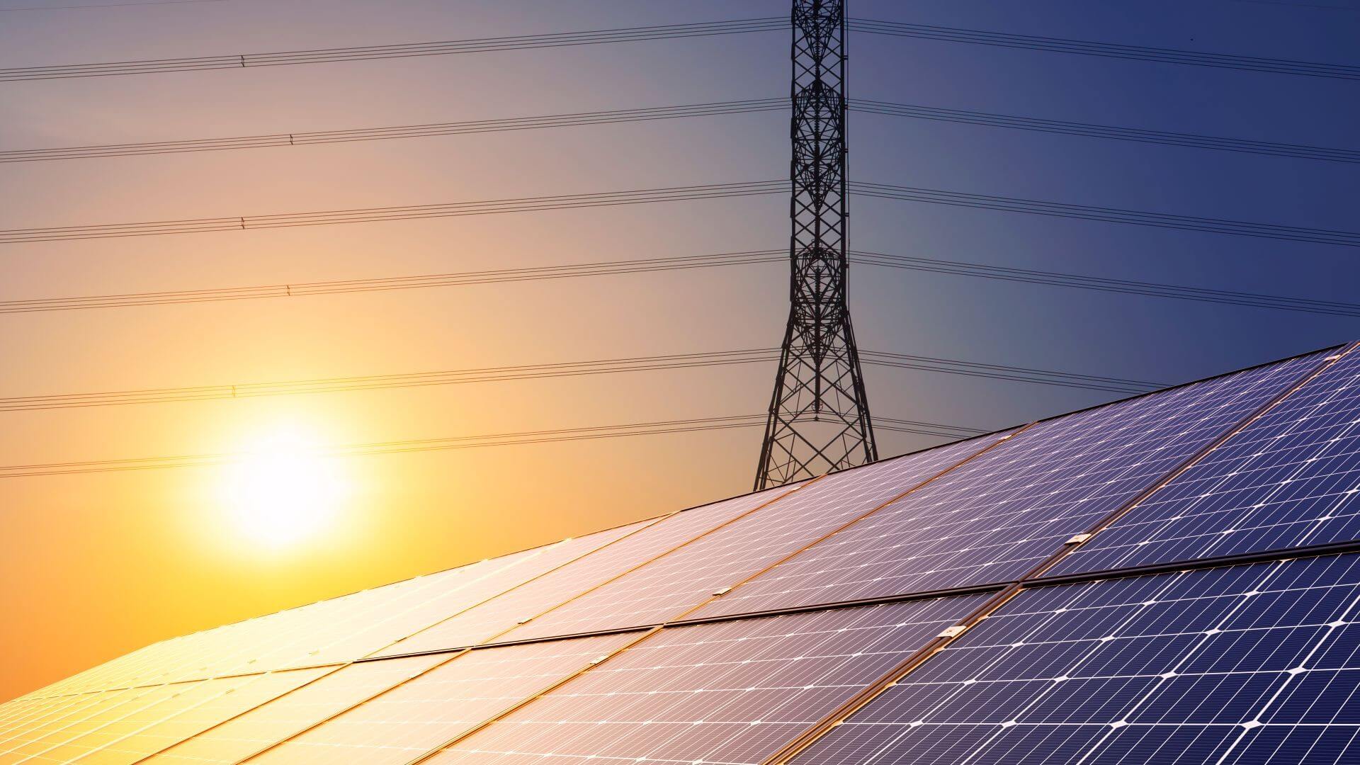 Close up of solar panels with an electricity pylon (side on) and transmission lines behind and sun in the sky